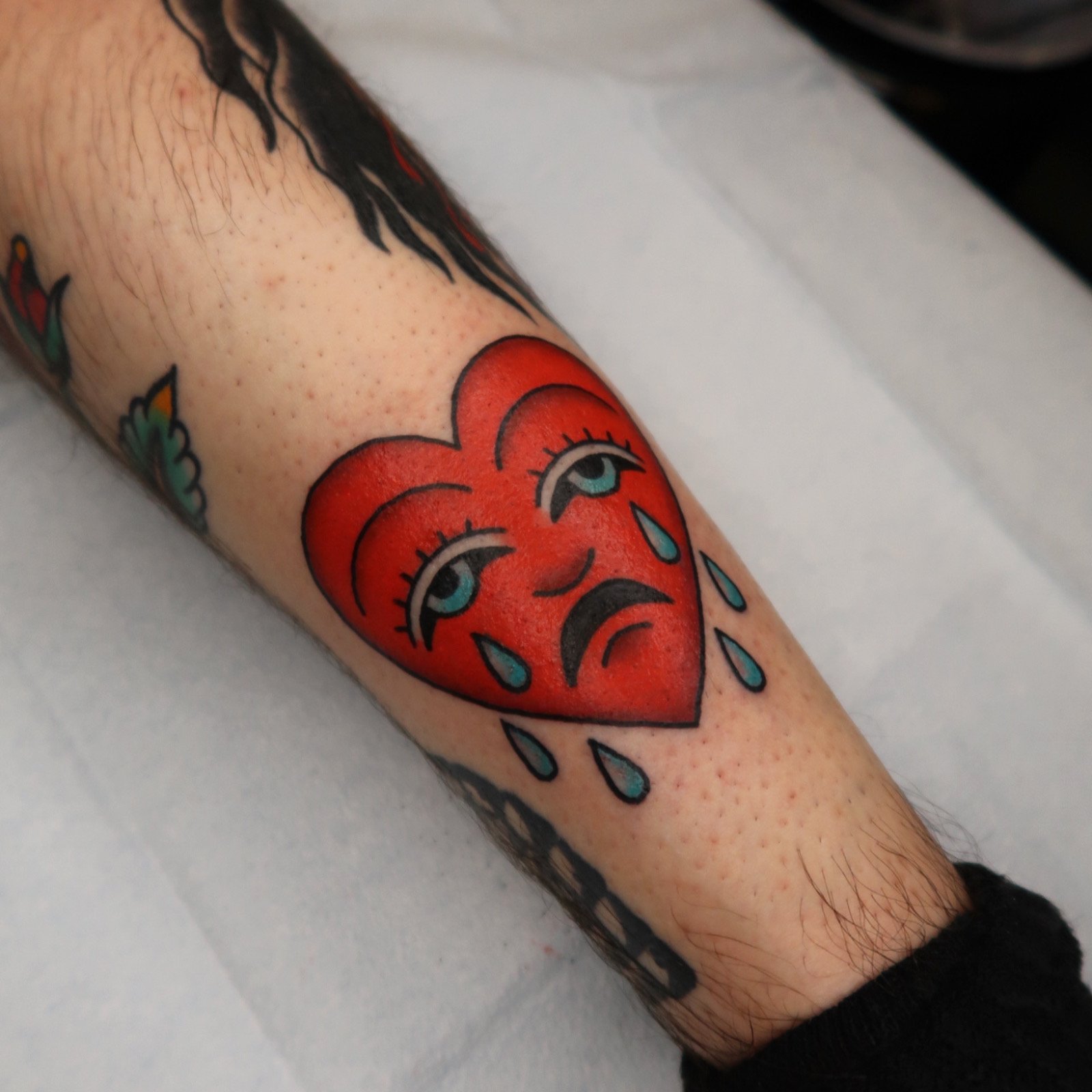 A colorful tattoo of a crying red heart with blue tears and eyes, on a person's leg.