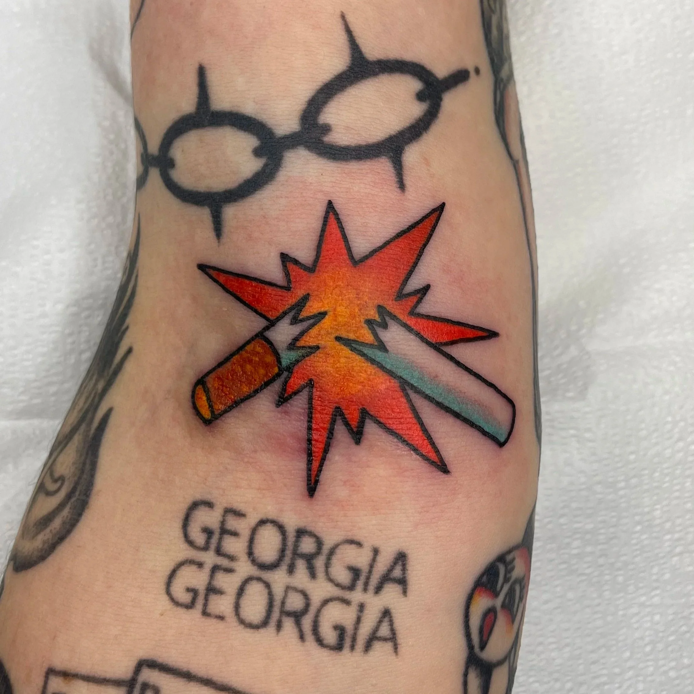 Tattoo featuring a black outline of a broken cigarette with an orange and yellow explosion and a rainbow-colored flame, along with black text listing Georgia counties.
