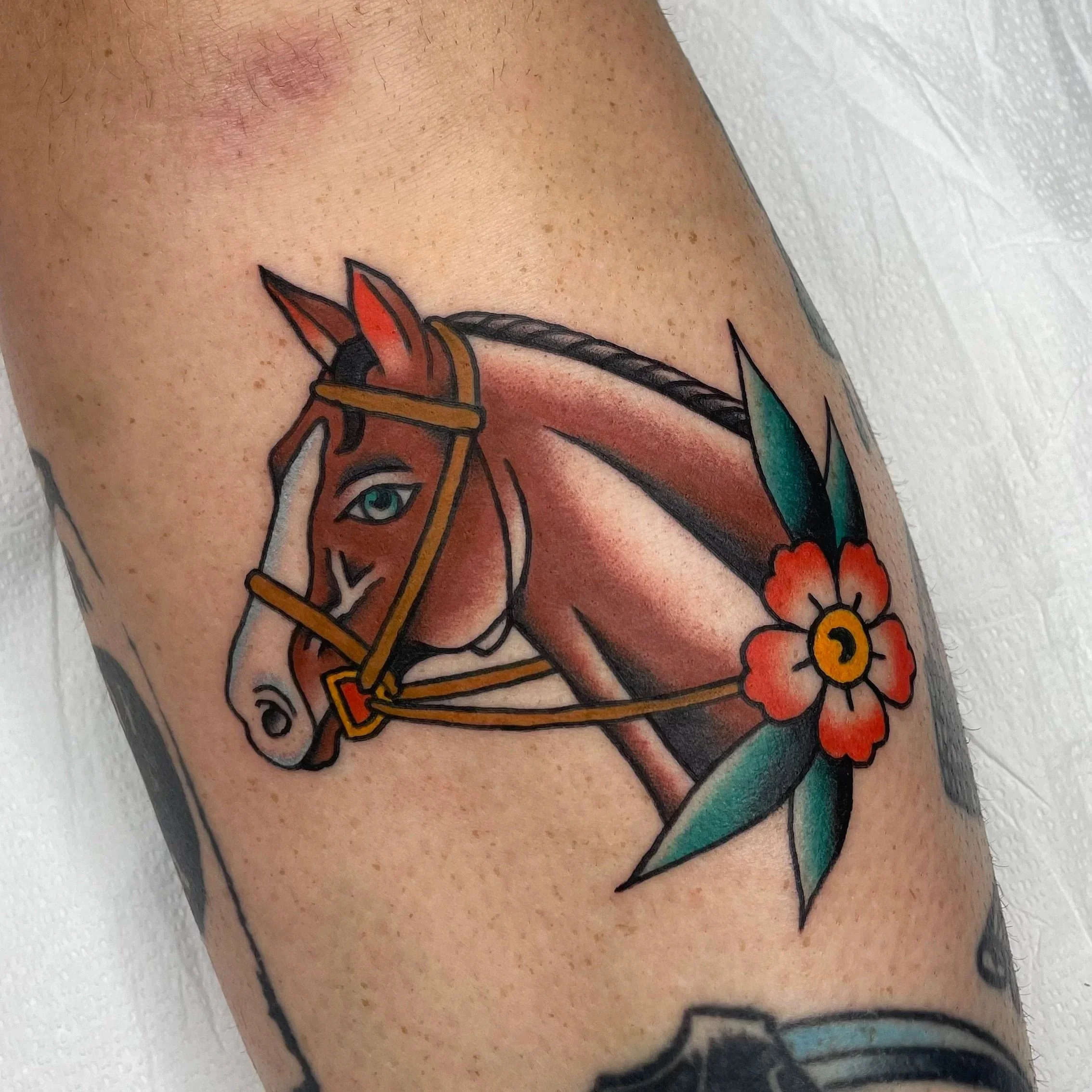 Tattoo of a horse with a colorful mane on a person's arm, with visible freckles and part of a sleeve tattoo.
