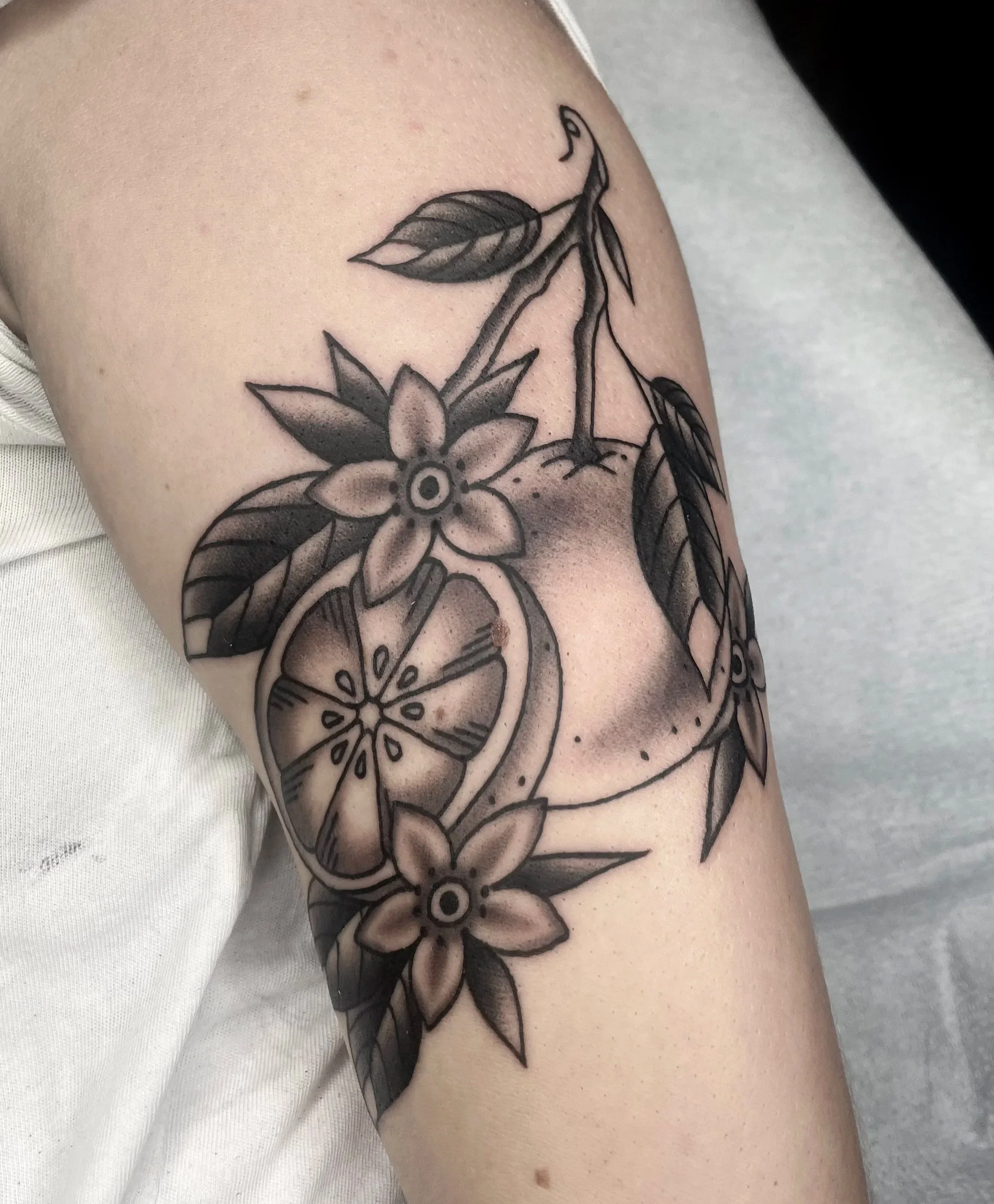 Black and gray floral tattoo on person's upper arm, featuring a flower, leaves, and a orange slice.