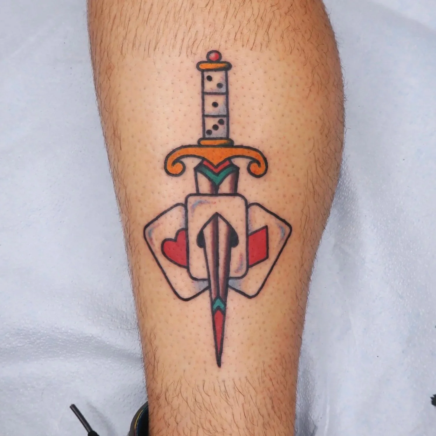 Tattoo of a sword with a heart-shaped guard, and a dotted design on the blade, on a person's leg.