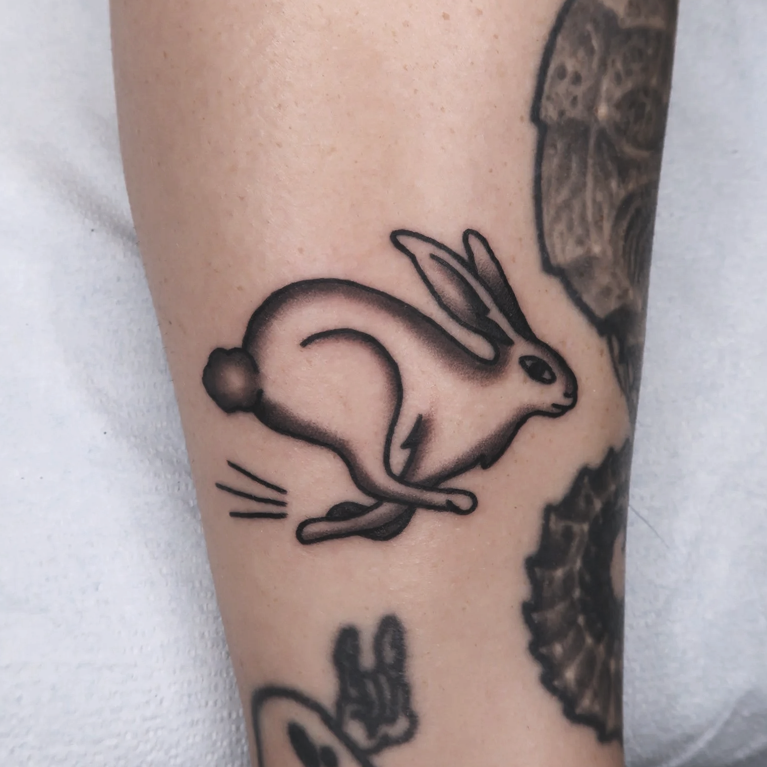 A tattoo of a stylized rabbit with a rounded body, large ears, a tail, and a small face, outlined in black ink on a person's leg.