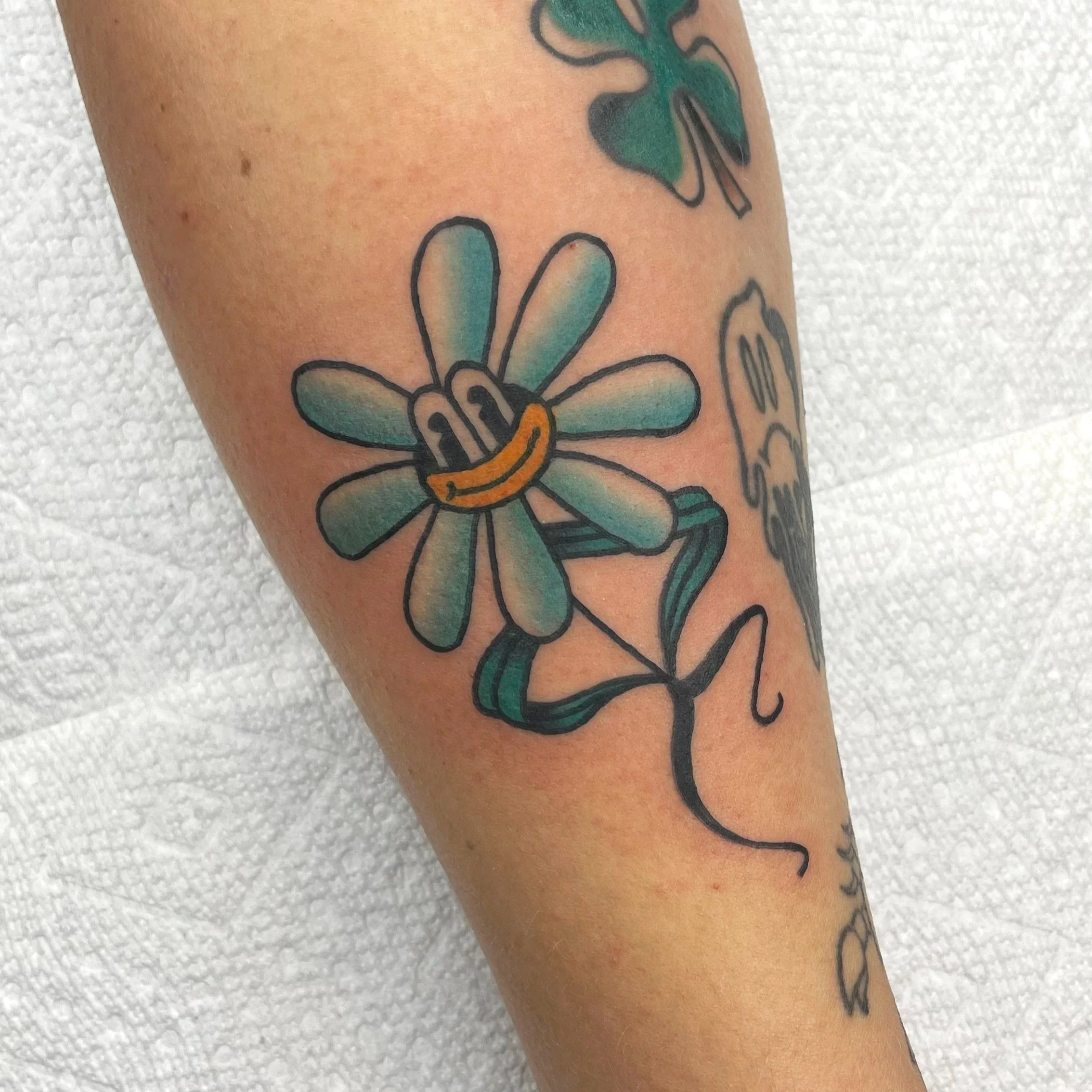 Colorful tattoo of a cheerful flower with a yellow and black striped body, petals with a gradient of blue and green, and a big smile, on a person's arm against a textured white background.