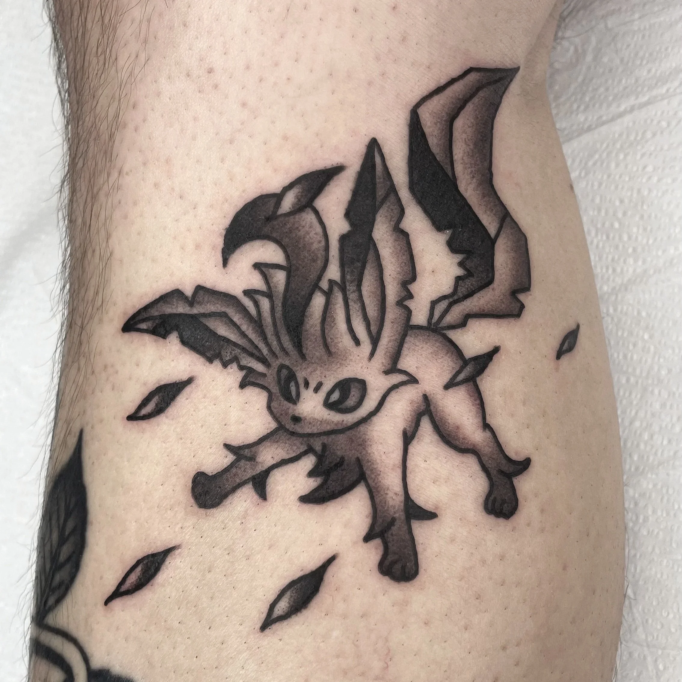 Close-up of a human skin with black ink tattoo of a pokemon design, showing parts of petals and leaves.