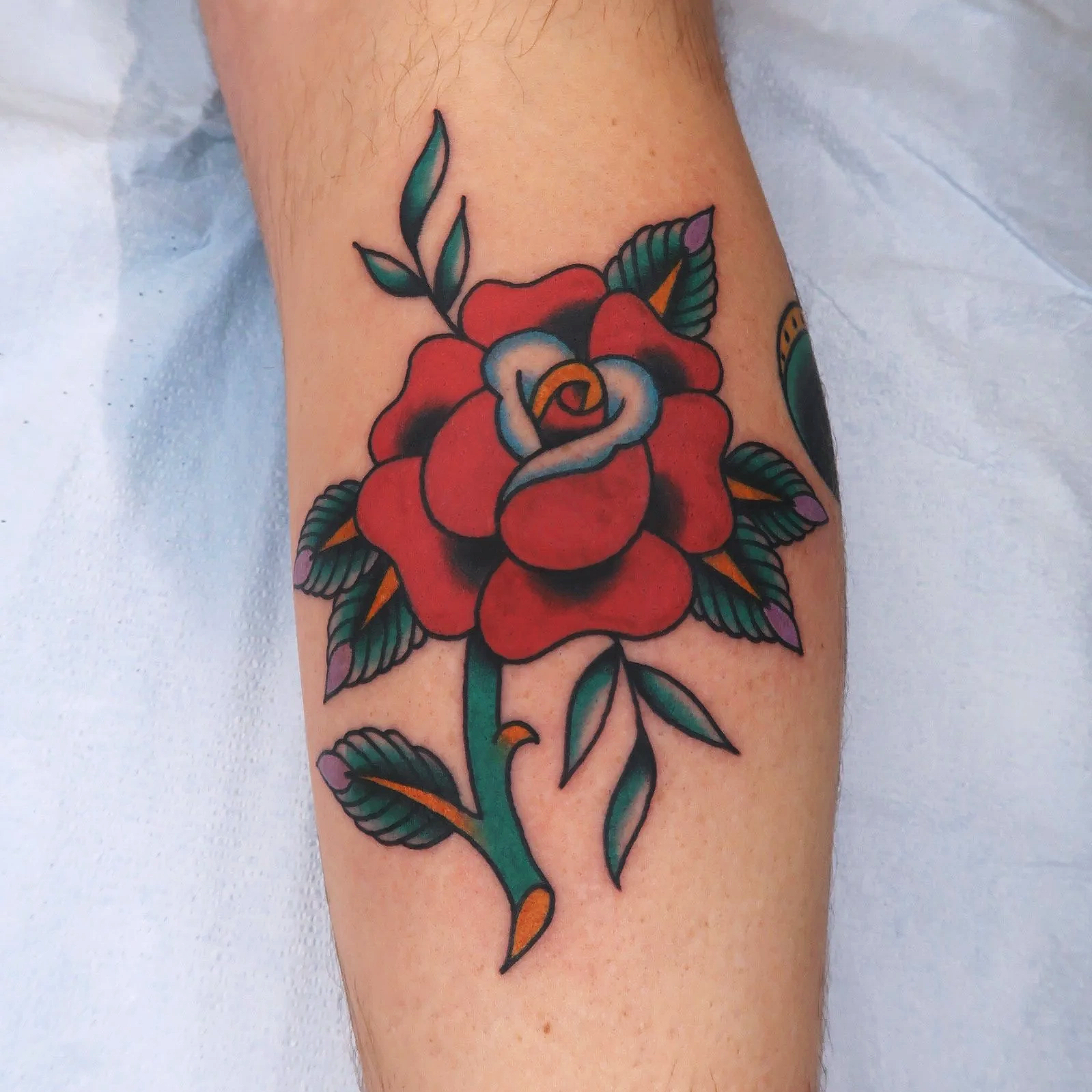 Colorful tattoo of a red rose with green leaves on a person's arm.
