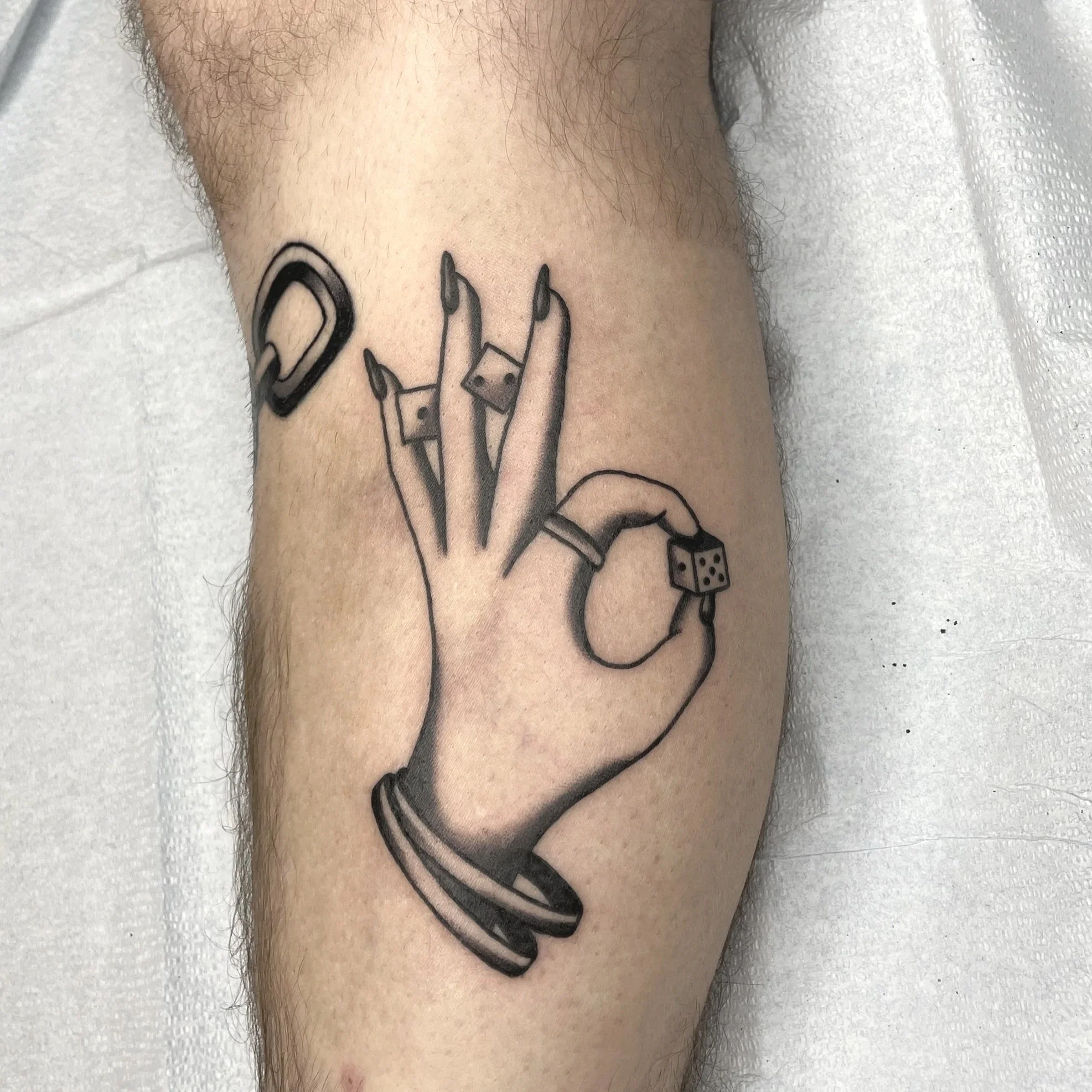 Tattoo of a finger with a ring, holding a pair of dice, on a person's skin.