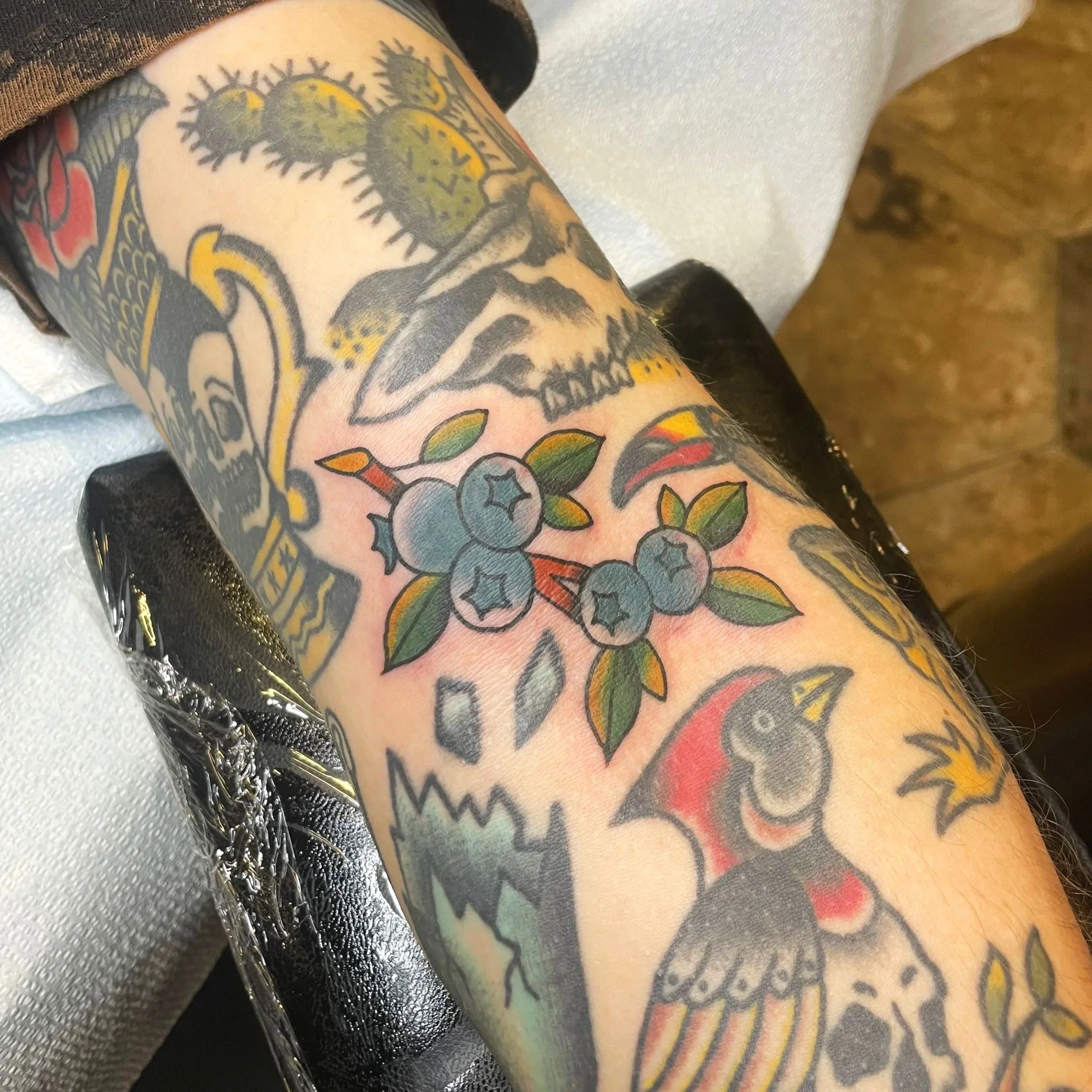 Close-up of a person's arm with colorful tattoo artwork, including a yellow skull with dark outlining, green leaves, and other vibrant designs.