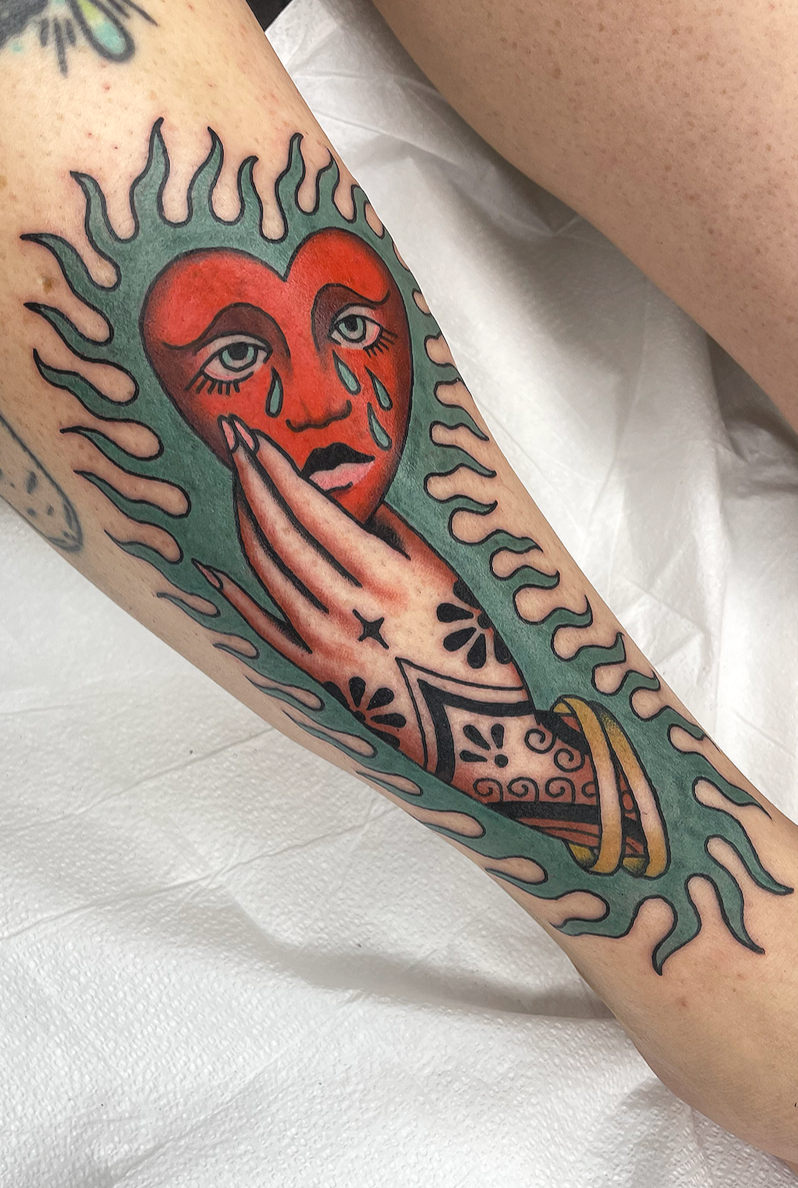 Tattoo of a crying heart with a face, tears, and a hand touching its face, surrounded by a green flame-like border.