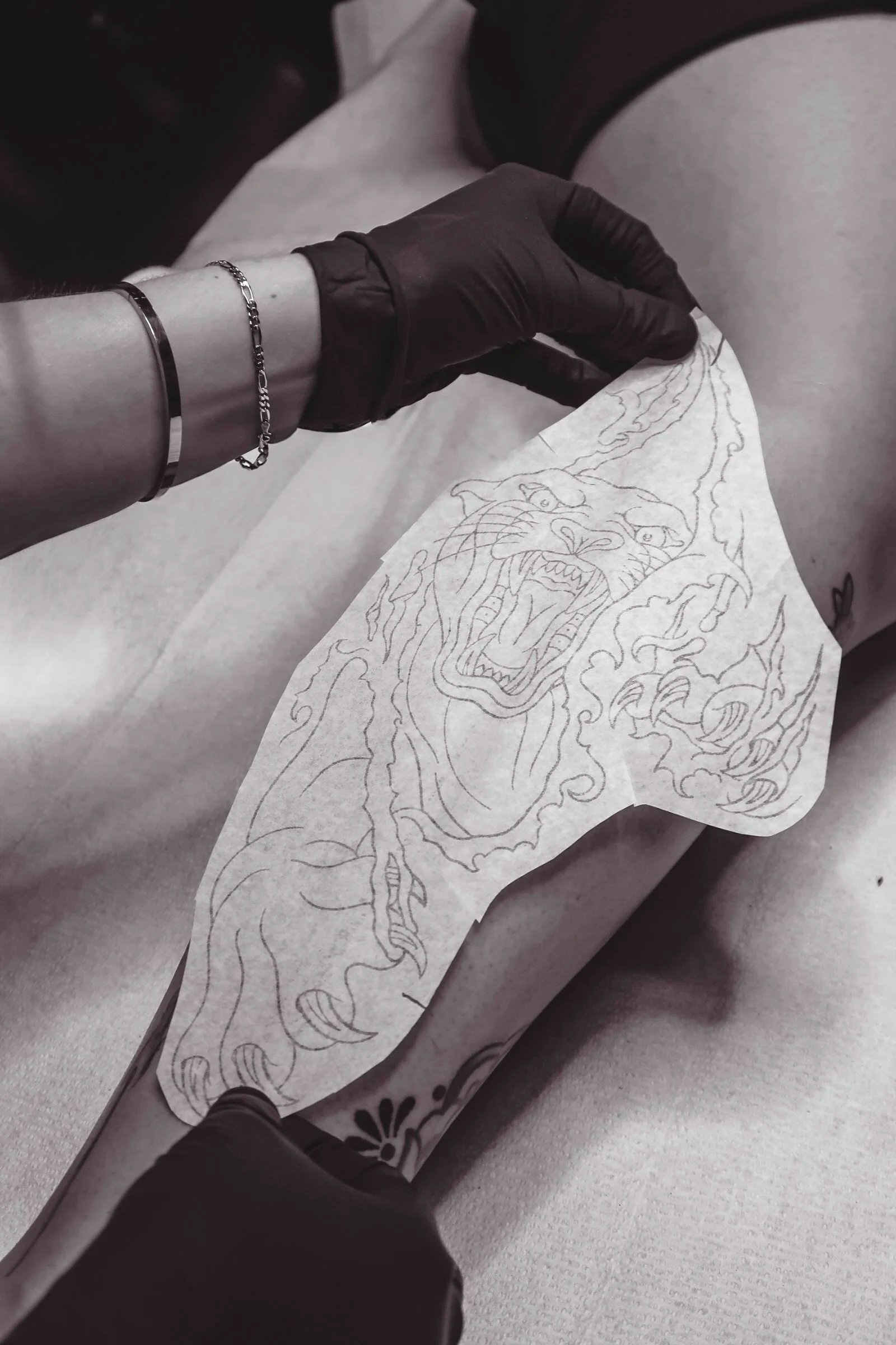 A person wearing black gloves is placing a tattoo stencil of a panther. The person is wearing bracelets on the wrist and the background is blurred.