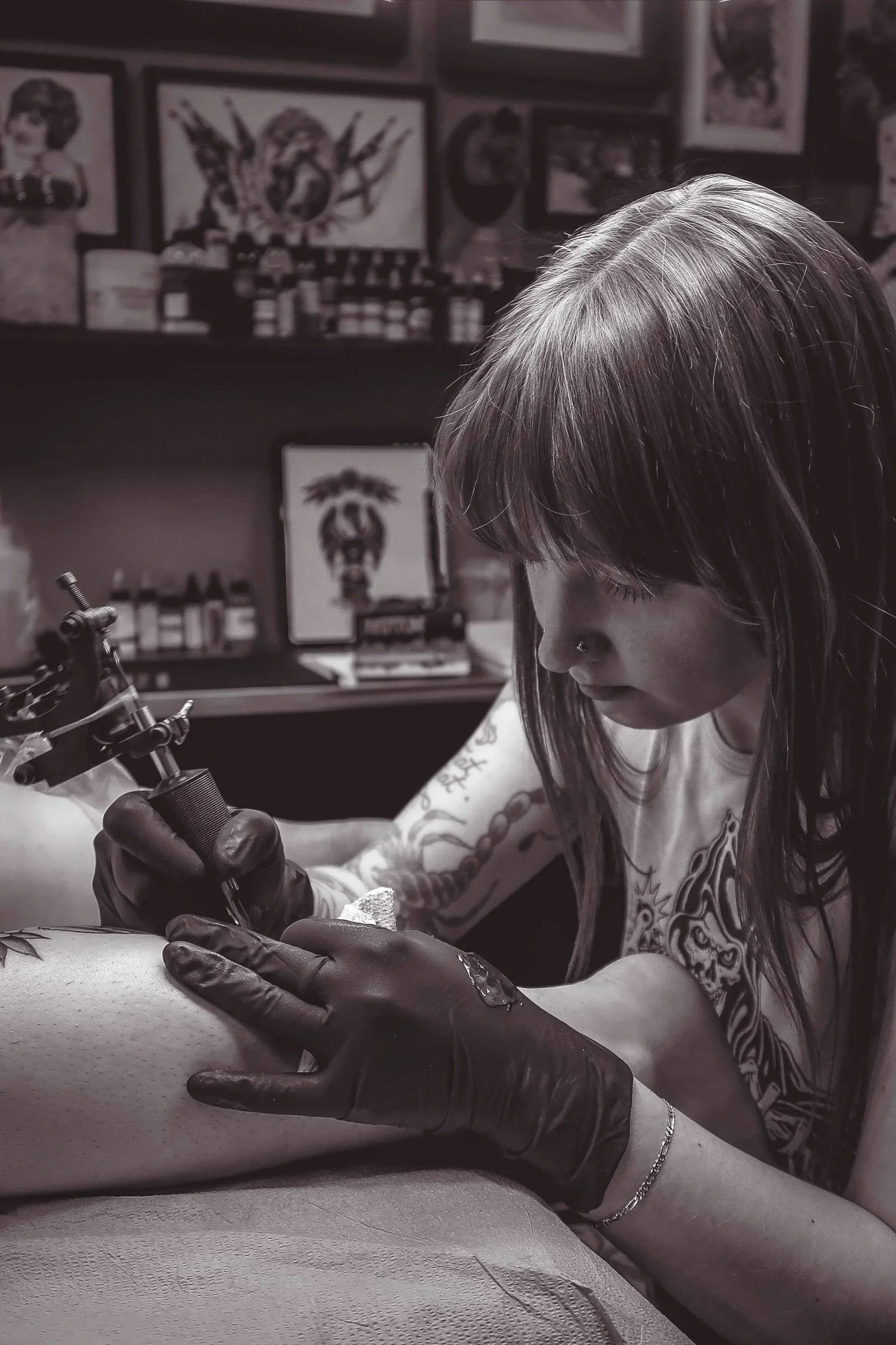 A person with long hair and tattoos on their arms tattooing a person's skin in a tattoo studio, with framed artwork and tattoo supplies in the background.