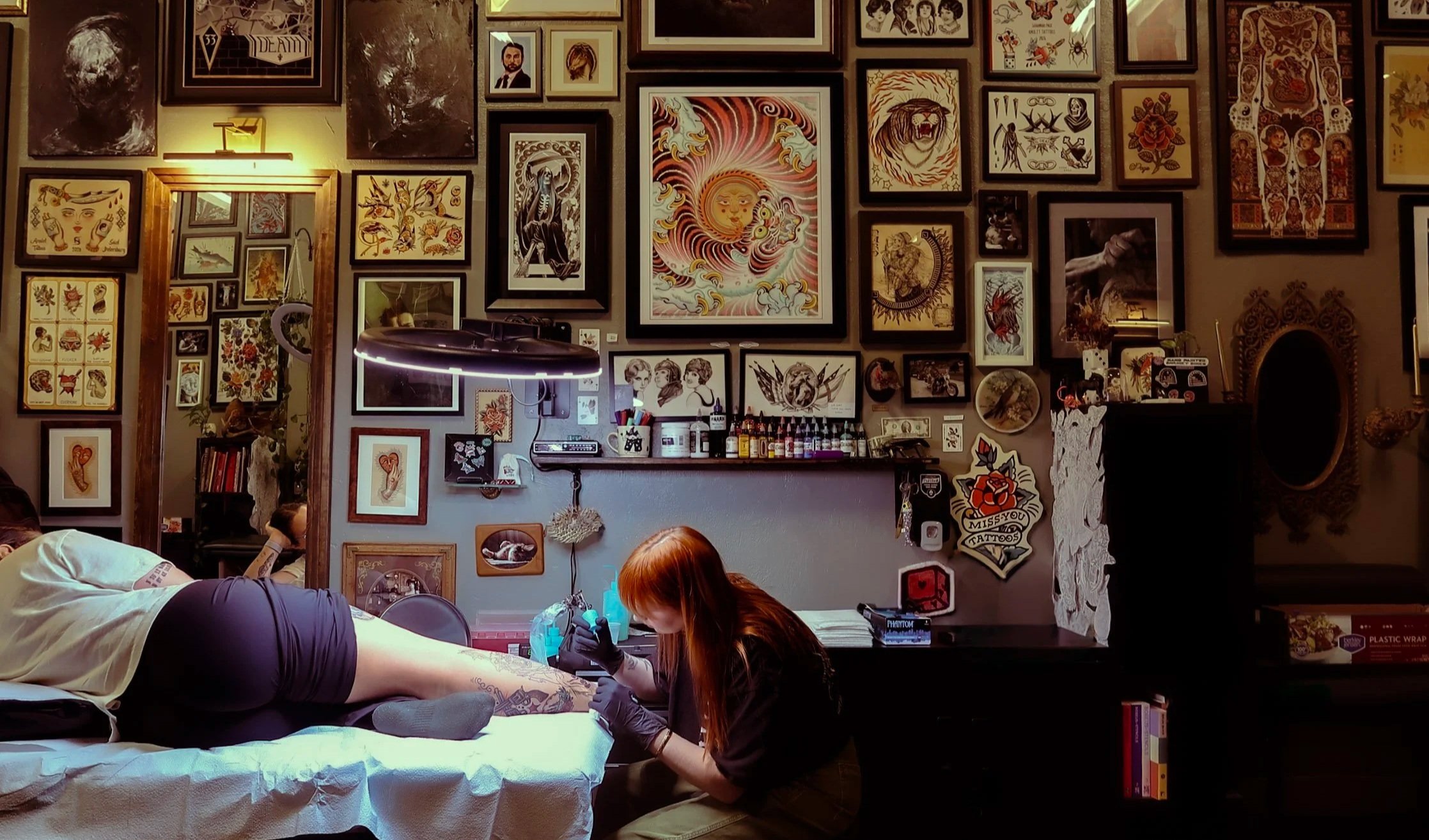 A tattoo artist working on a client's leg in a tattoo studio decorated with various framed artwork and tattoos on the wall.