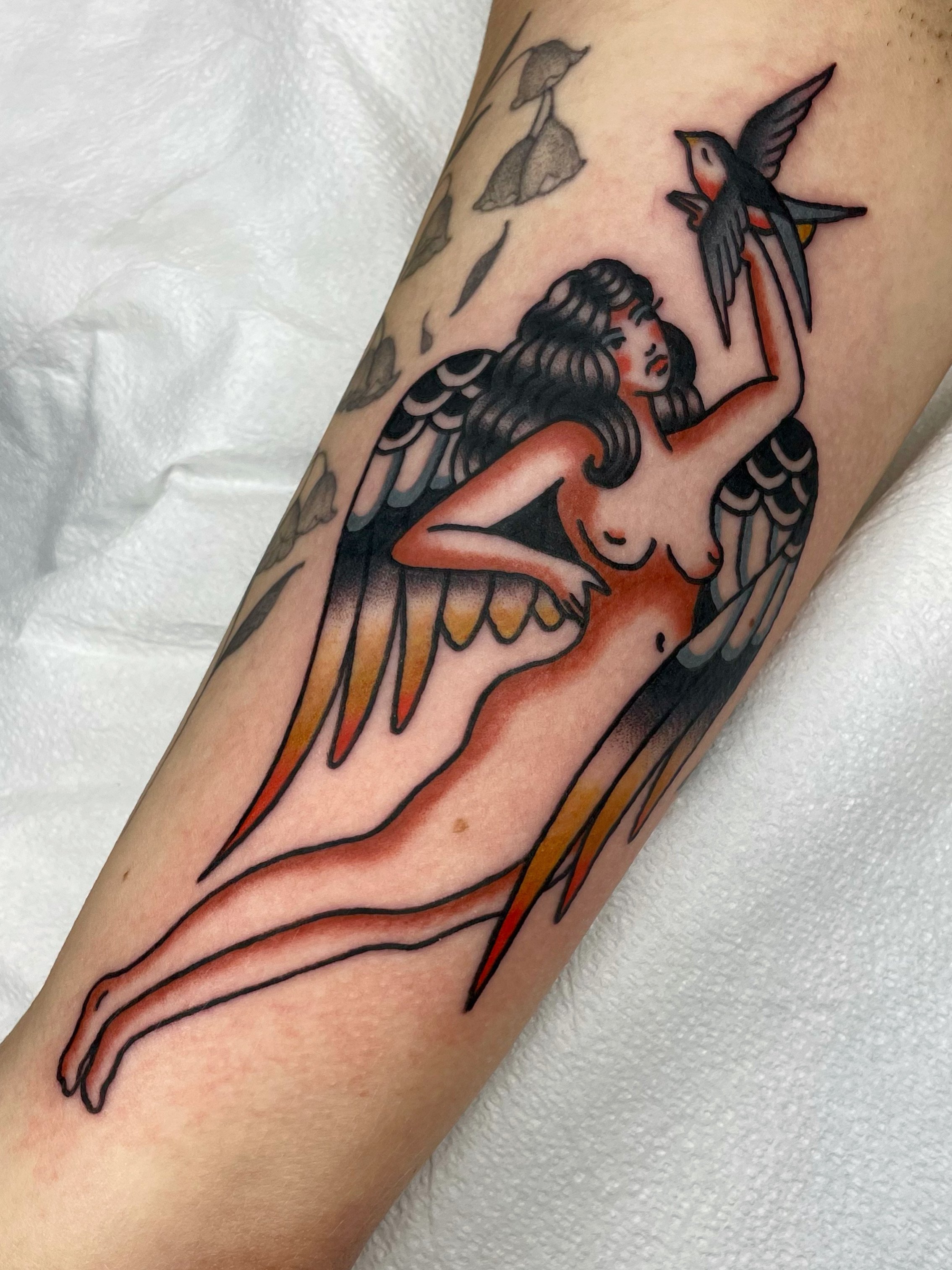 Tattoo of a woman with wings holding a flying bird on her finger.