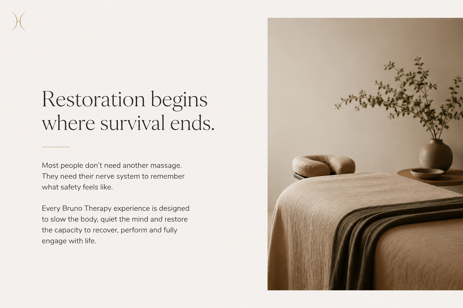 A minimalist massage therapy room with a neatly made bed covered in beige linen, a sculpted ottoman, and a side table with a large vase holding green foliage, all set against a neutral wall.