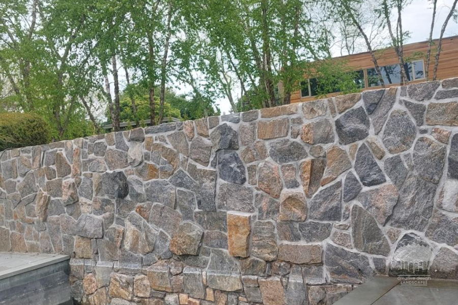 Natural fieldstone wall in varied gray and tan tones with trees behind it
