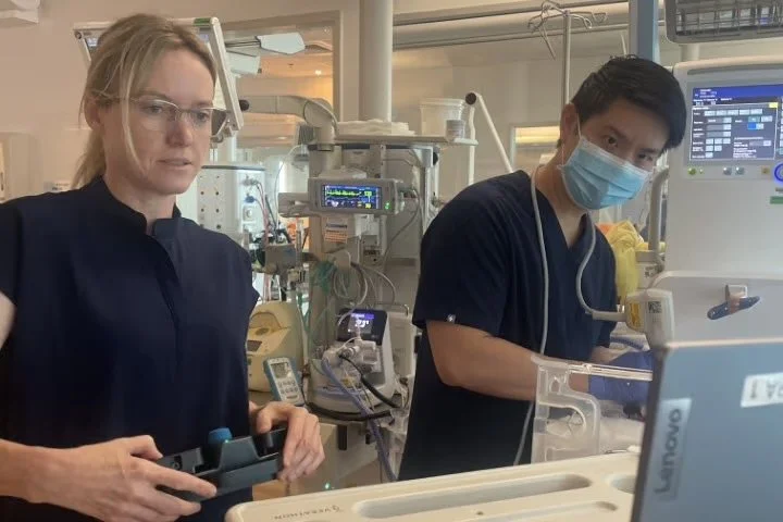 Two healthcare workers in a hospital Intensive Care Unit, wearing scrubs and a protective mask, operating medical equipment.
