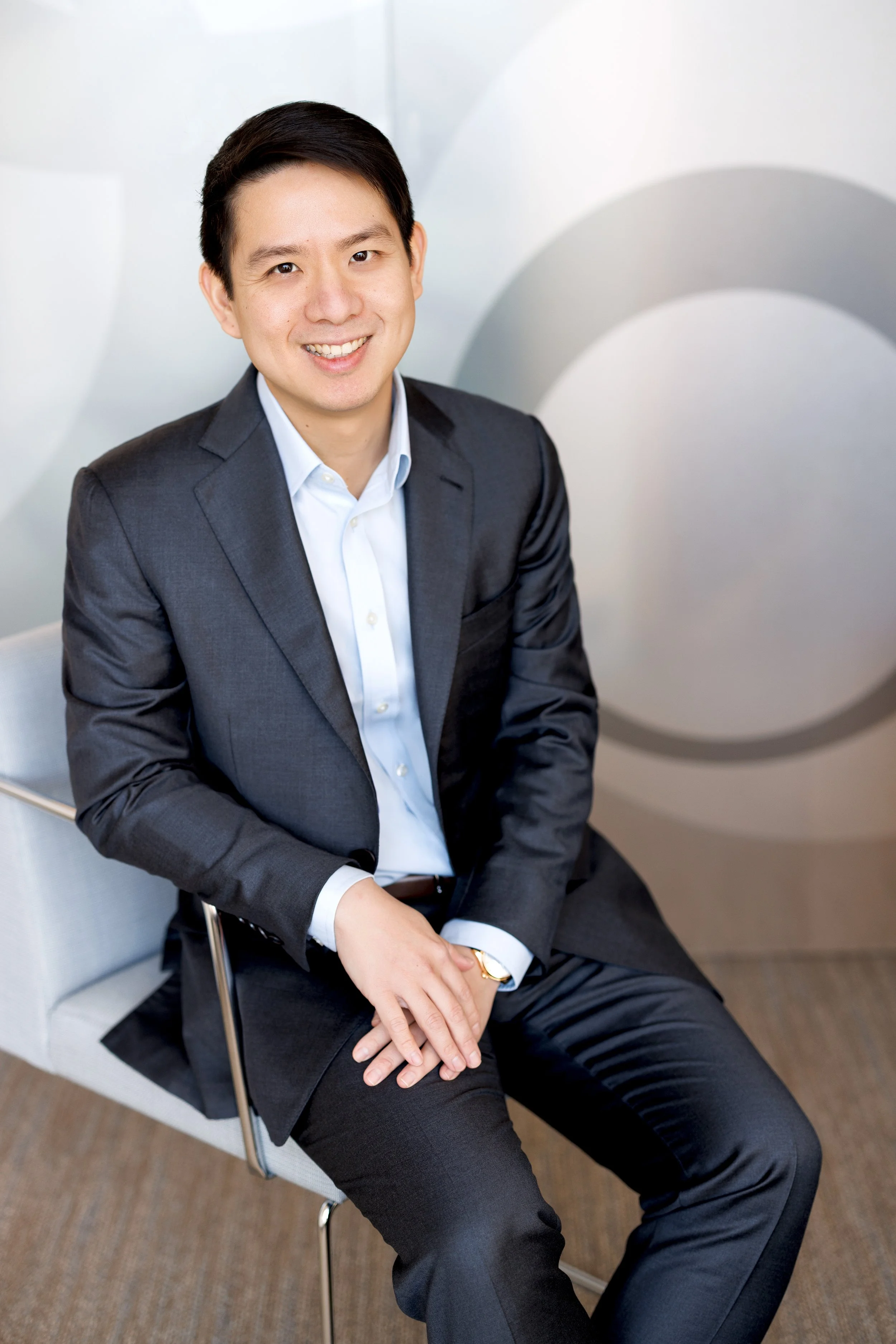 A man in a dark gray suit and white shirt sitting on a white chair in an indoor office environment, smiling at the camera.
