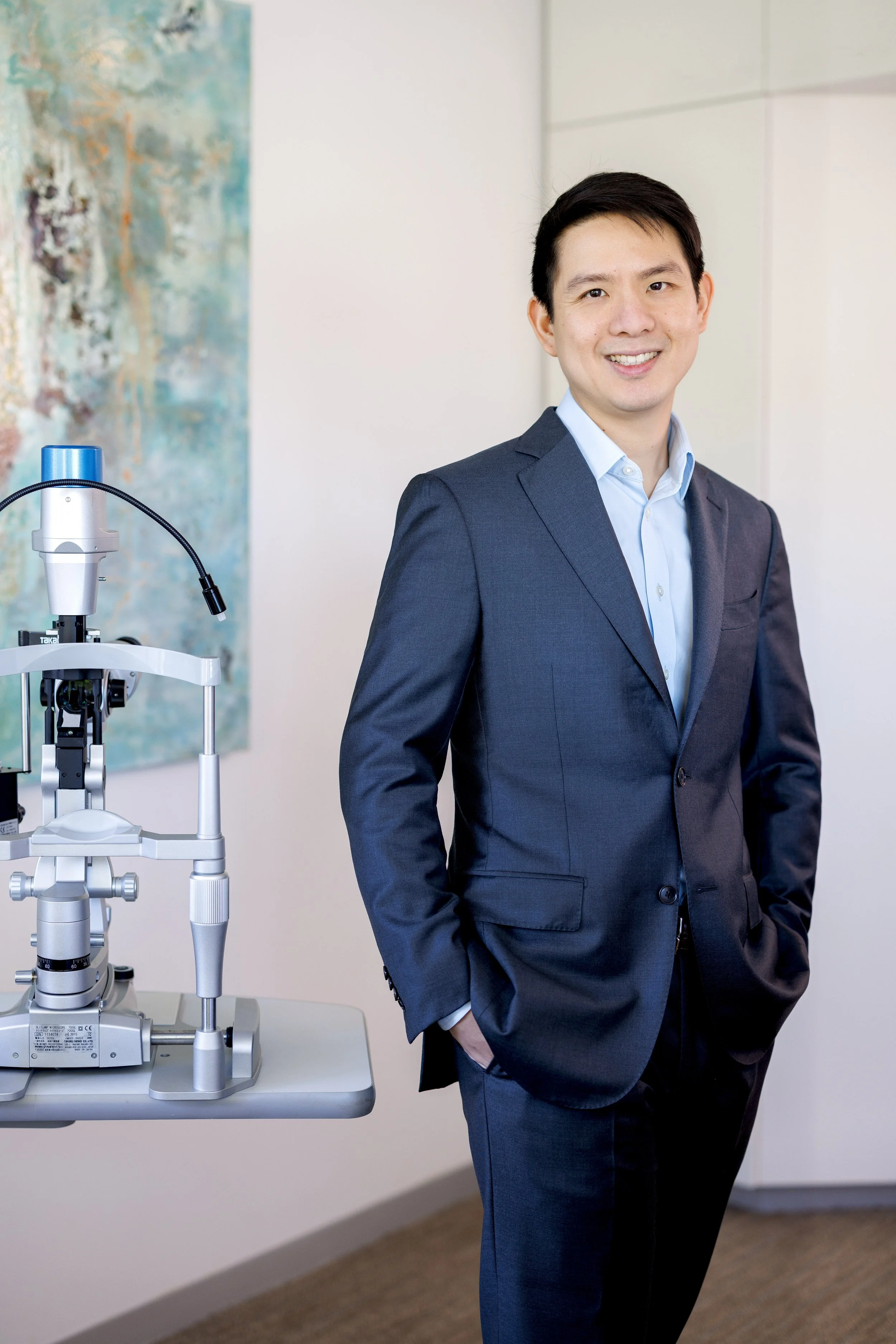 A man in a blue suit standing confidently with one hand in his pocket, smiling in a modern office or medical setting with an ophthalmology microscope nearby.