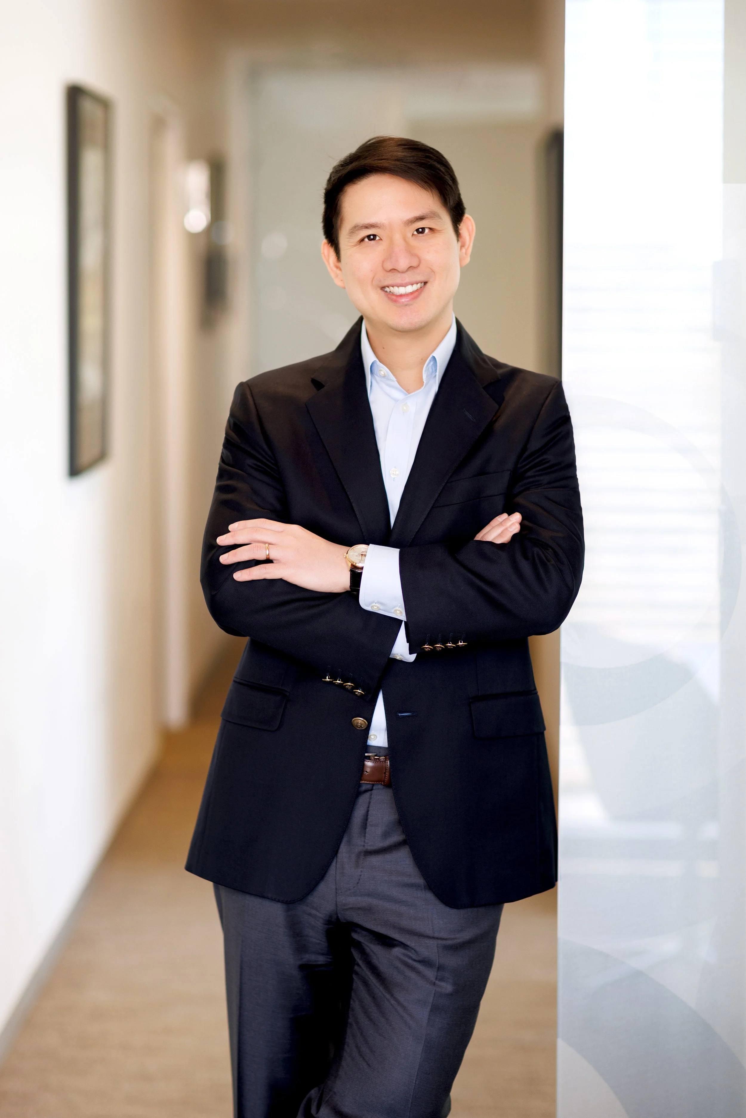 A smiling man in formal business attire standing with arms crossed in a hallway with framed artwork, near a window with natural light.