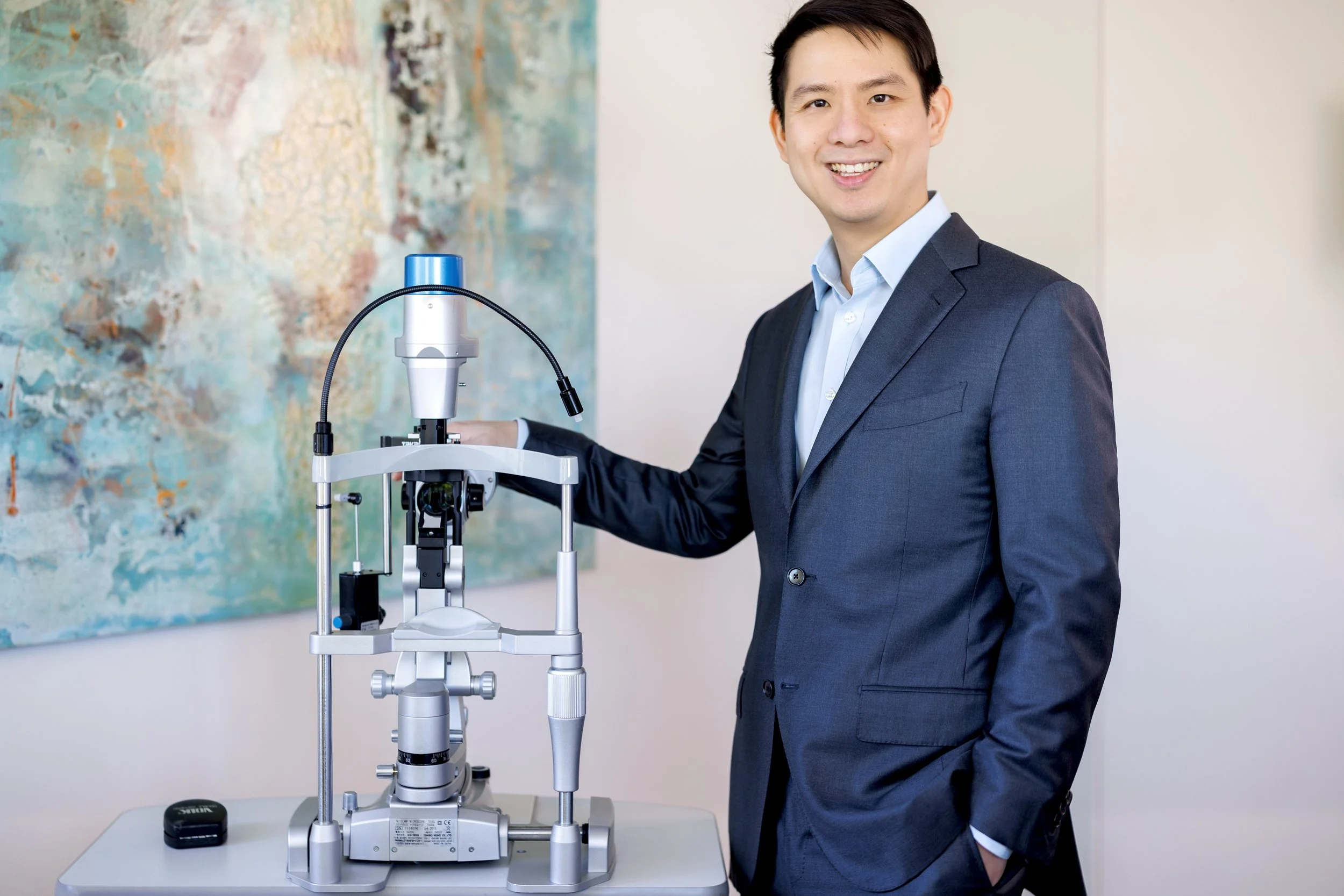 A man in a navy blue suit and light blue shirt smiling while standing next to a microscope on a table in a room with abstract artwork on the wall.