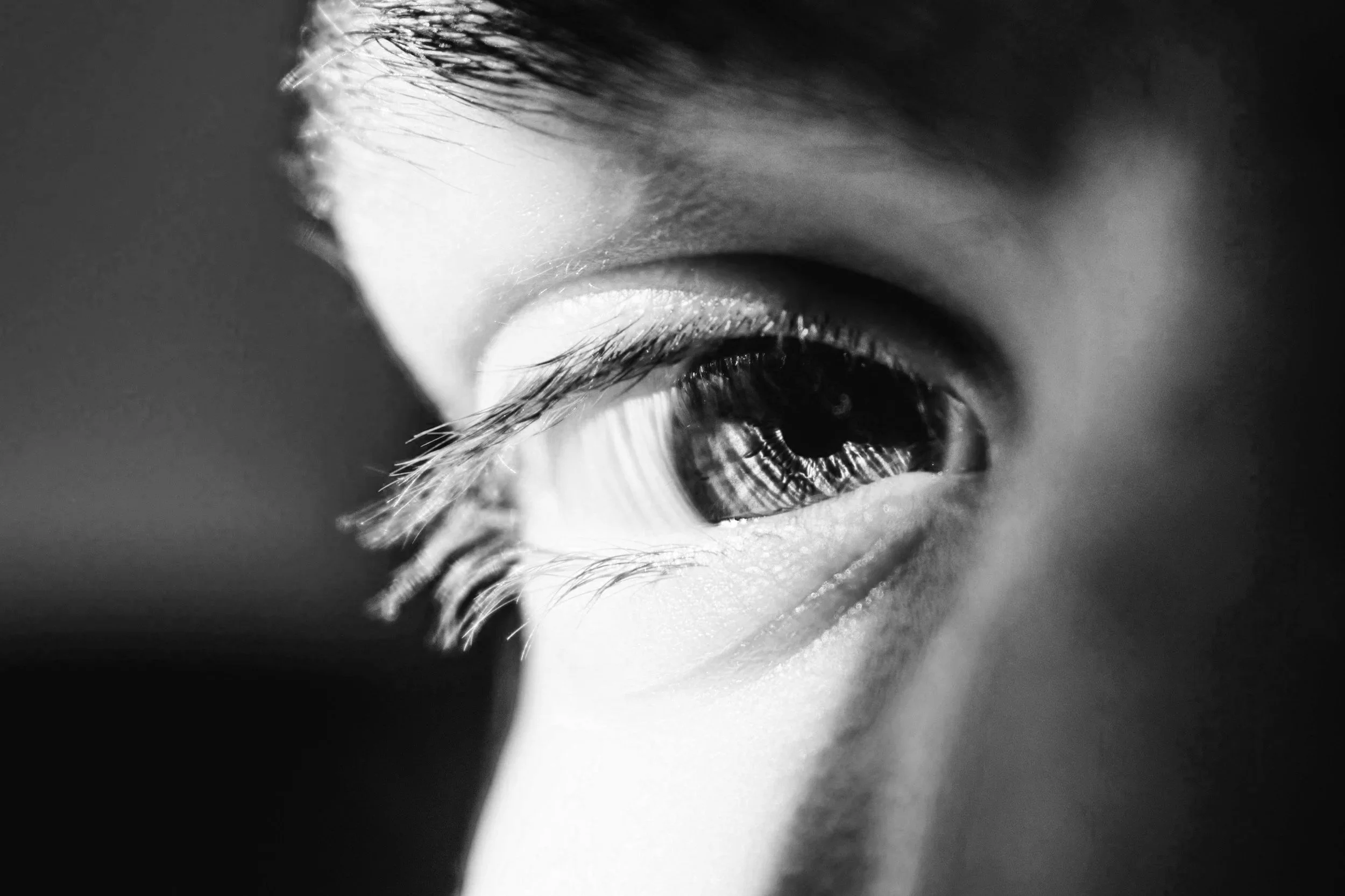 Close-up black and white photograph of a human eye, showing detailed eyelashes and skin texture.