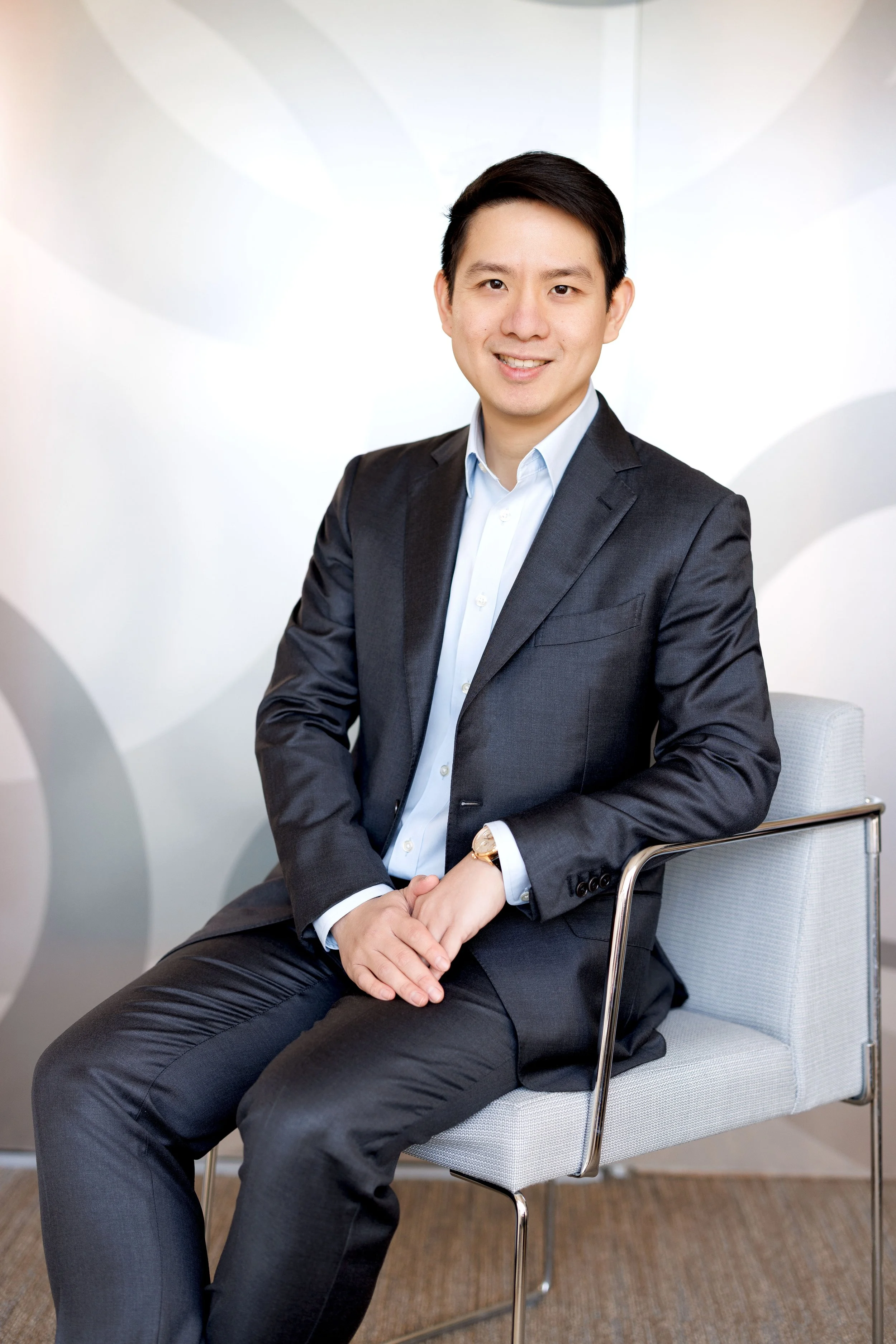 A young man in a dark suit and light blue shirt sitting on a modern gray chair with metal armrests, smiling at the camera, in a well-lit indoor setting with a white background featuring abstract light leather shapes.