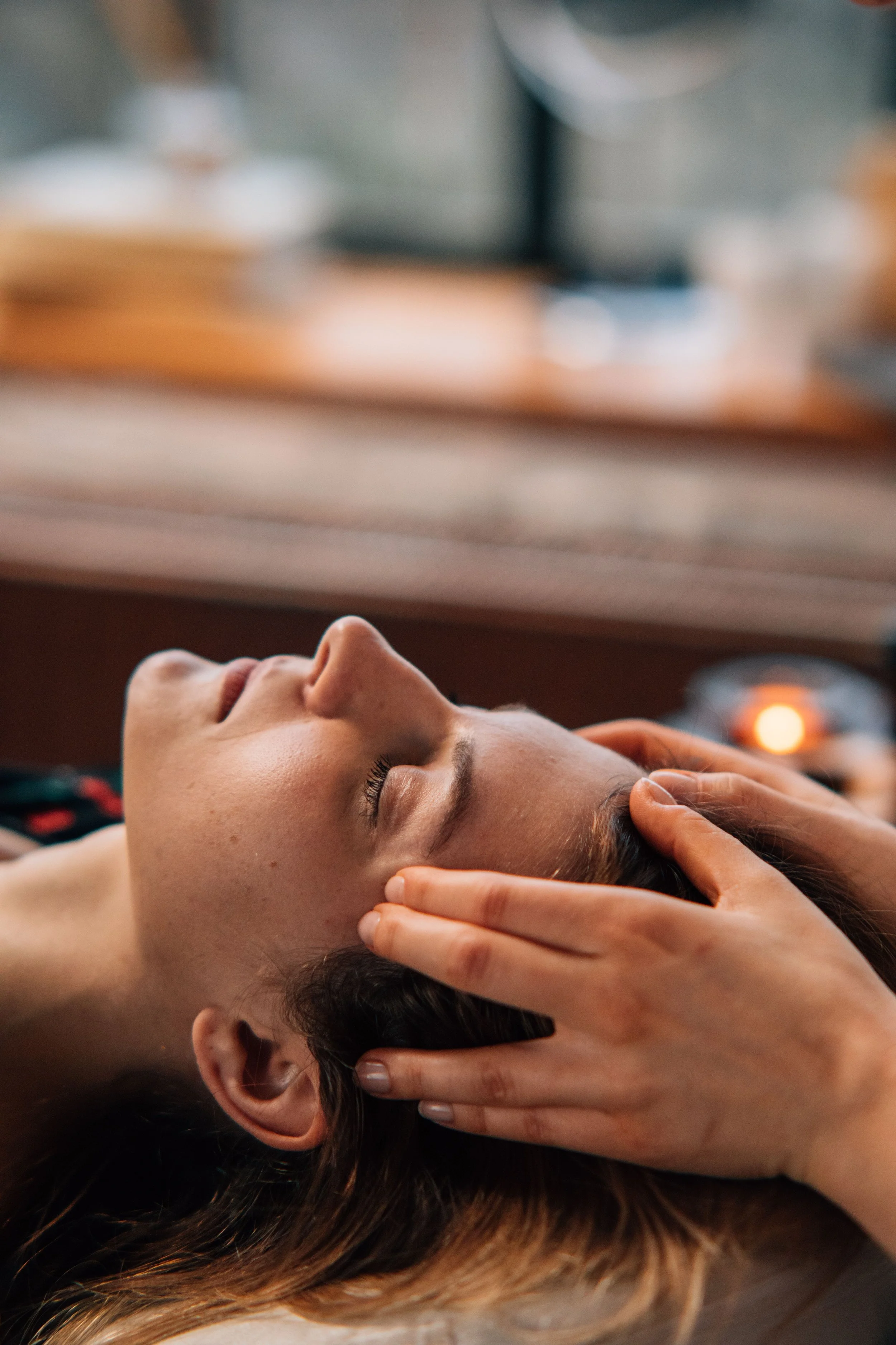 A person is lying down with eyes closed, receiving a massage or facial treatment. The person's head is supported by someone else's hands, and there is a soft, warm ambiance suggested by the blurred background and warm lighting.