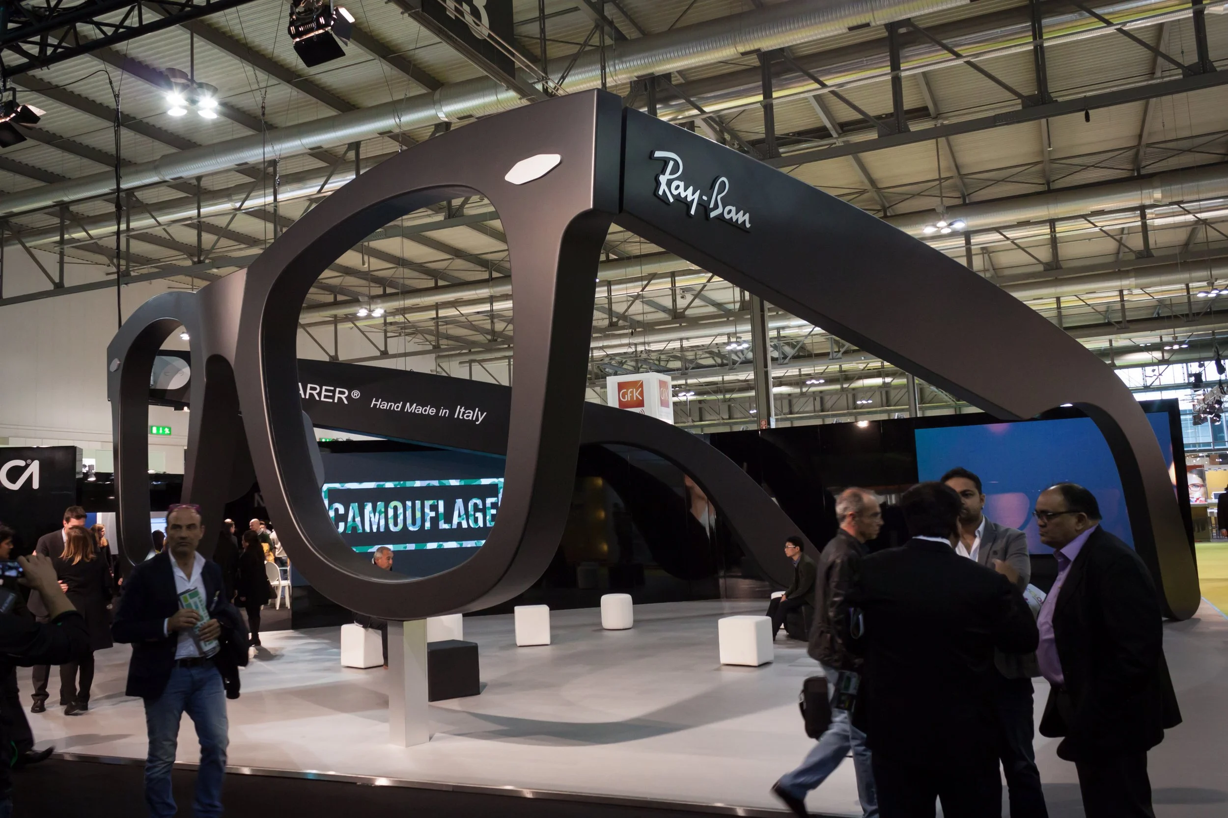 Trade show booth with large black frame resembling a camera lens, with the brand name 'Ray-Ban' written on top. People are walking and engaging around the booth in a large conference hall.