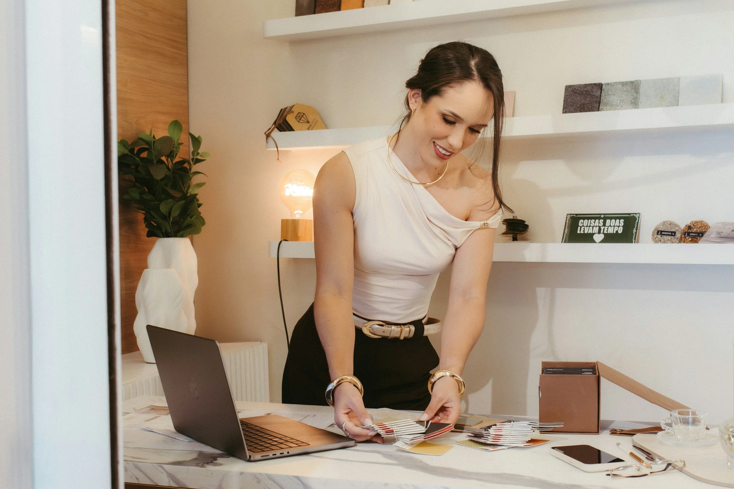 A woman in a white top and black skirt is working at a desk with color swatches. She smiles while organizing the swatches, with a laptop, notebooks, and various office supplies on the desk. Behind her are white shelves with decorative items and a sign that reads 'COISAS BOAS LEVAM TEMPO'.