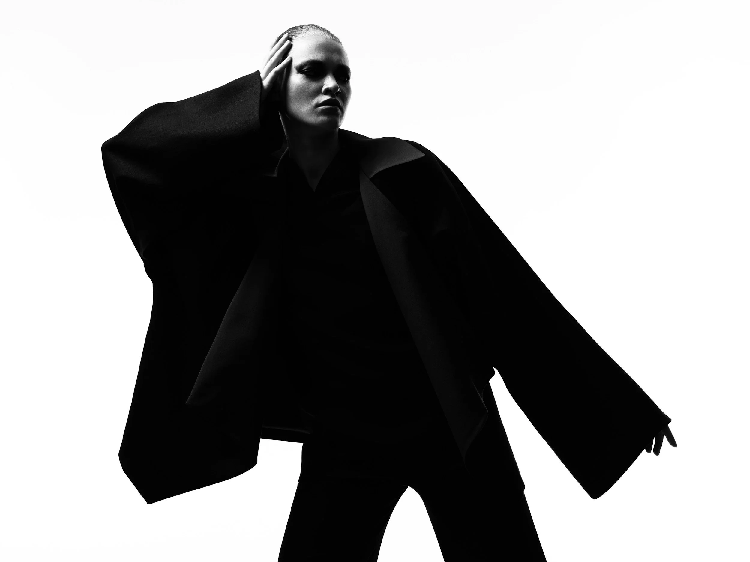 A black and white photo of a woman with platinum blonde hair, wearing a dark oversized blazer, posing with one hand on her head and looking serious against a plain white background.