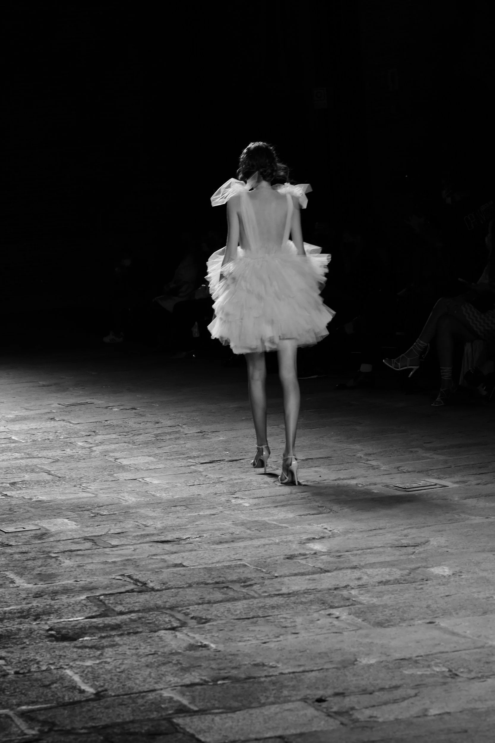 A woman wearing high heels and a layered, ruffled, backless dress walking on a cobblestone street at night.