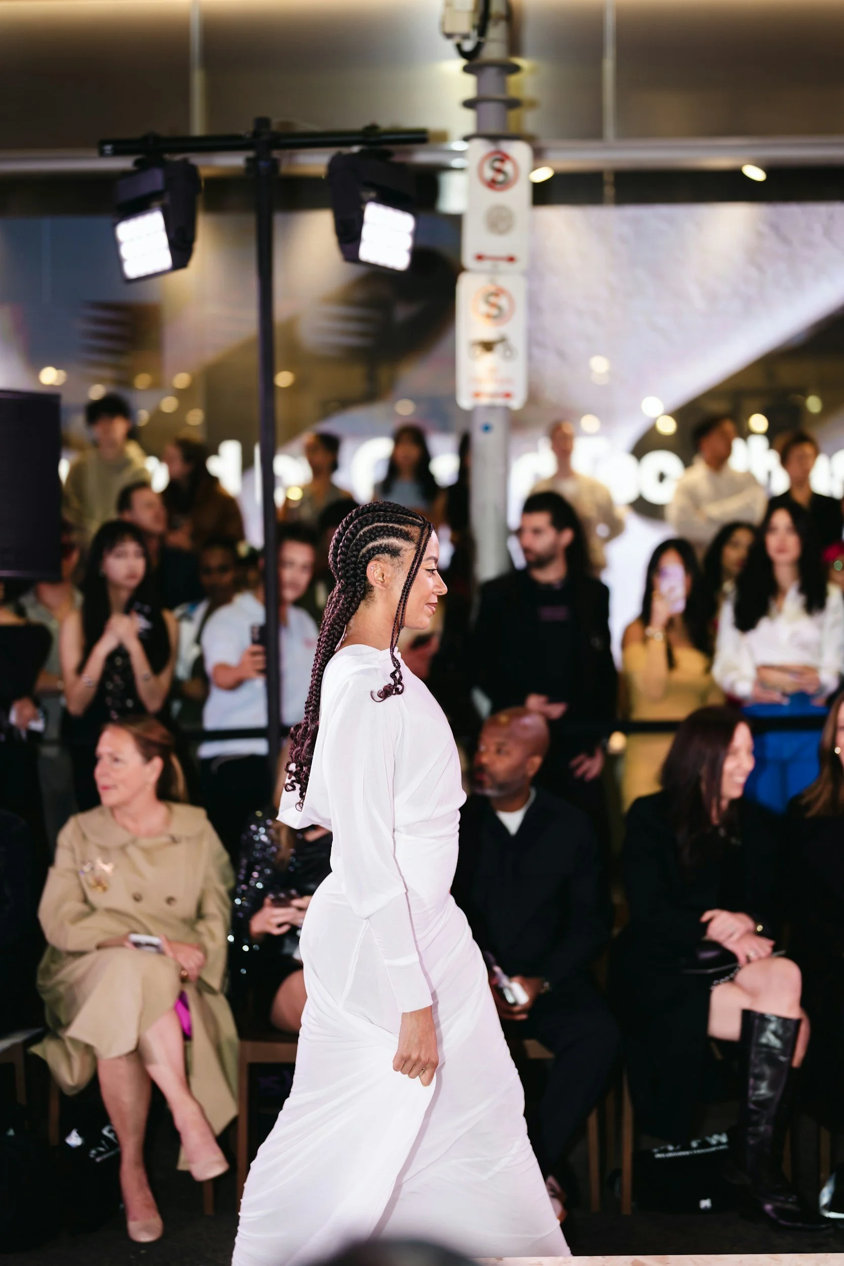 A woman with braided hair in a white outfit walking in front of an audience at a fashion show.