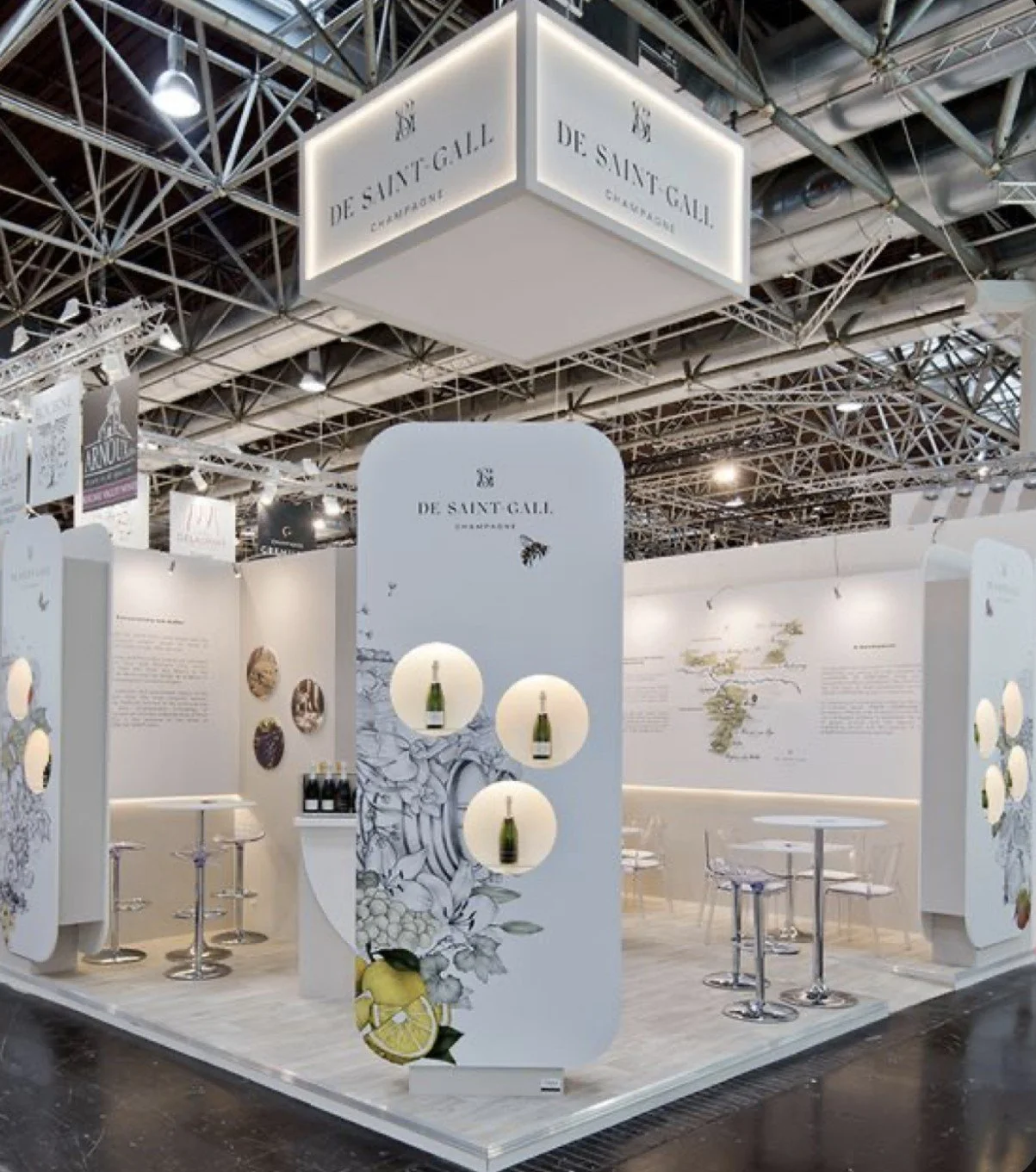Exhibit booth for De Saint Gall Champagne at a trade show or fair with white walls, round tables, and decorative bottles of champagne displayed on circular cutouts on a white panel.