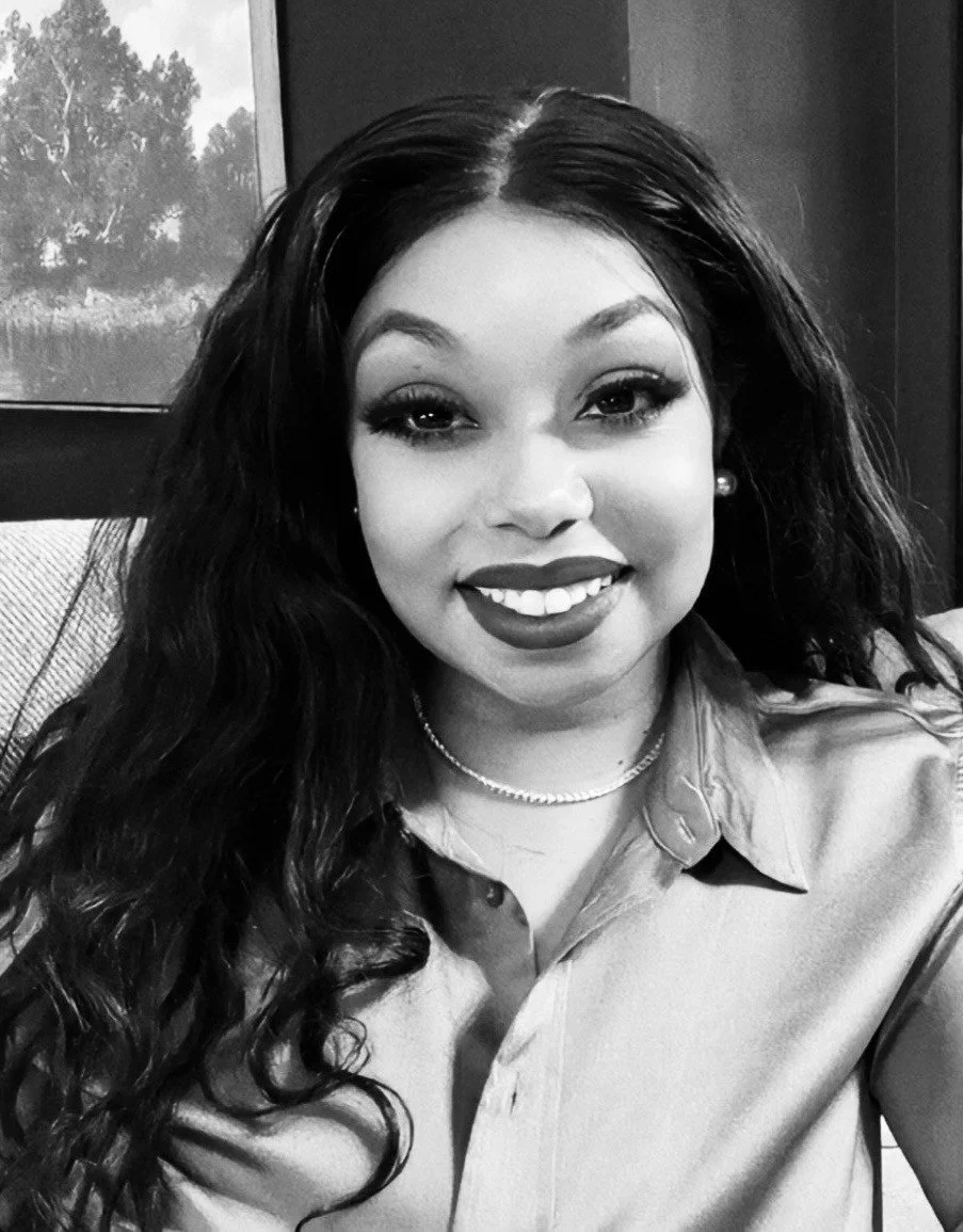 A woman with long curly hair, smiling, wearing a collared shirt, earrings, and a necklace, in a black-and-white photo.