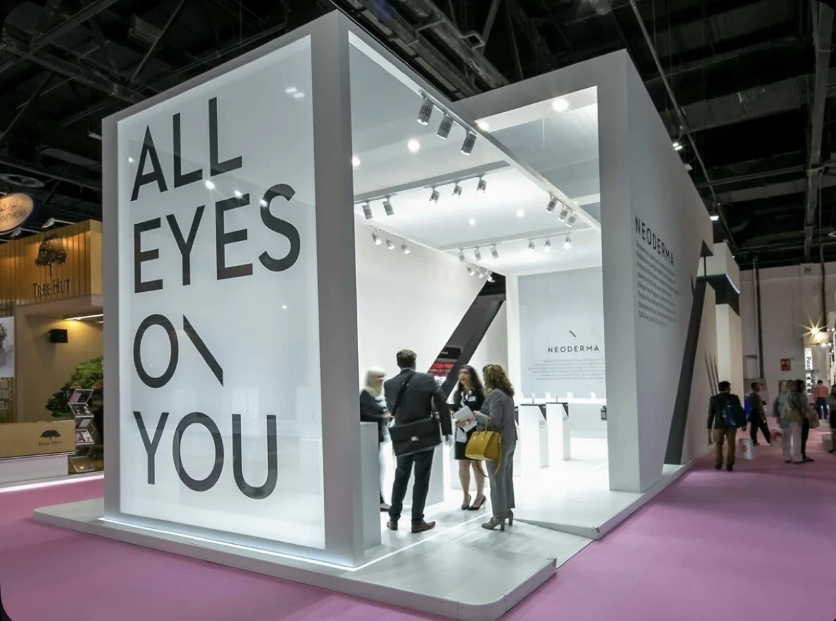 Trade show booth with large sign that says 'ALL EYES ON YOU' and smaller text about NeoDerma skincare products, with people engaging in conversation inside the booth.
