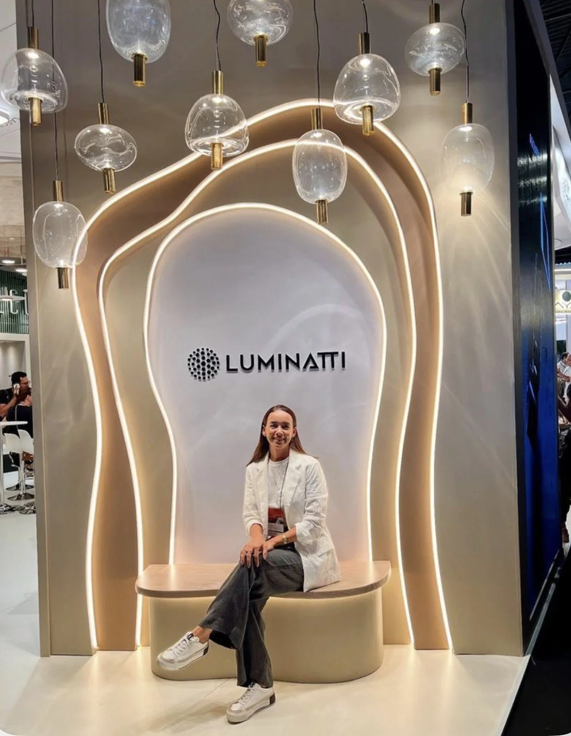 A woman sitting on a modern beige bench in front of a display with the Luminit logo, illuminated wavy structures, and hanging glass pendant lights in a stylish exhibition booth.