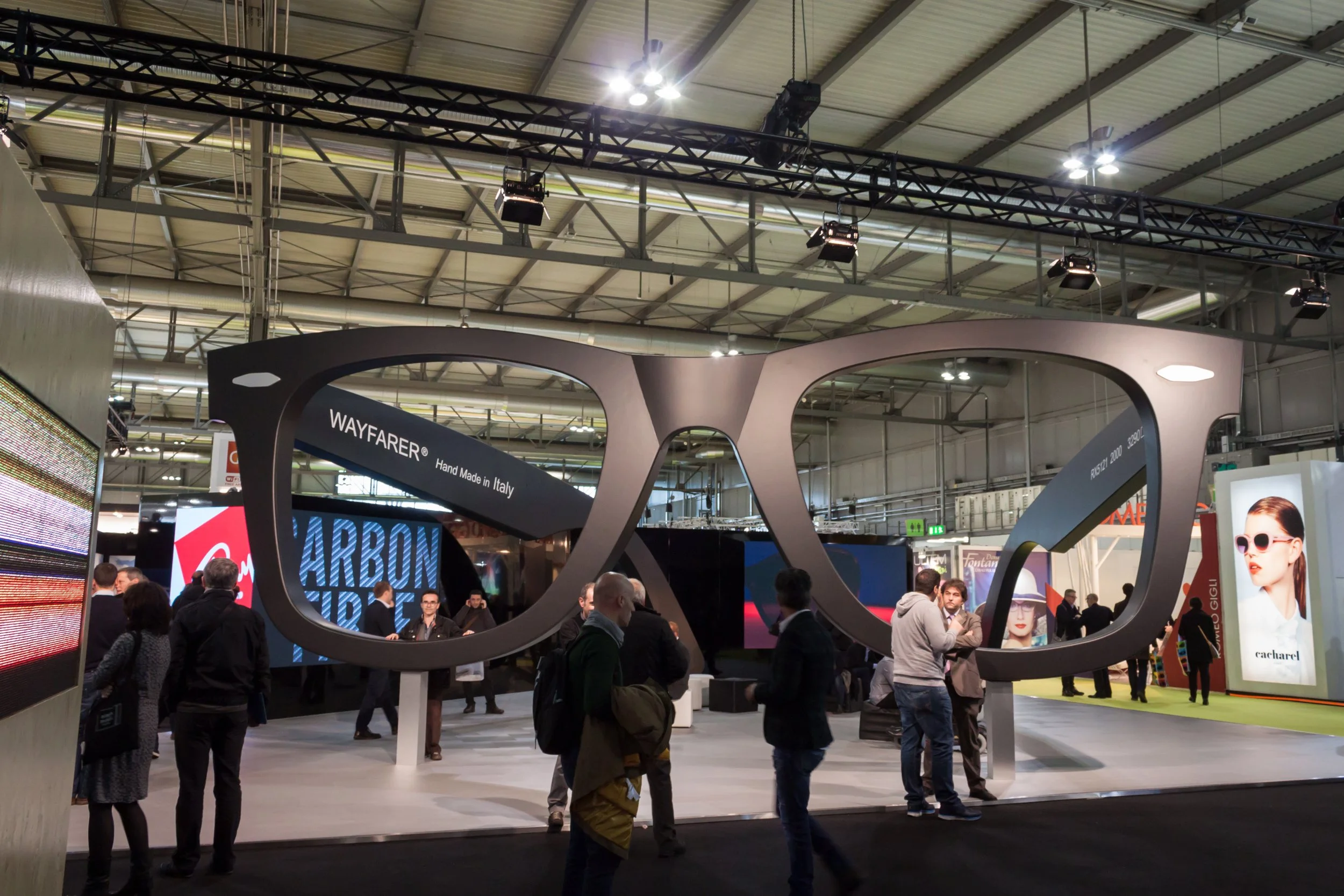 An indoor exhibition hall filled with people walking and standing around various displays and booths. Visible signage includes 'Carbon Fibre' and 'Wayfarer,' with a large digital screen displaying colorful visuals. The ceiling features industrial lighting and metal framework.