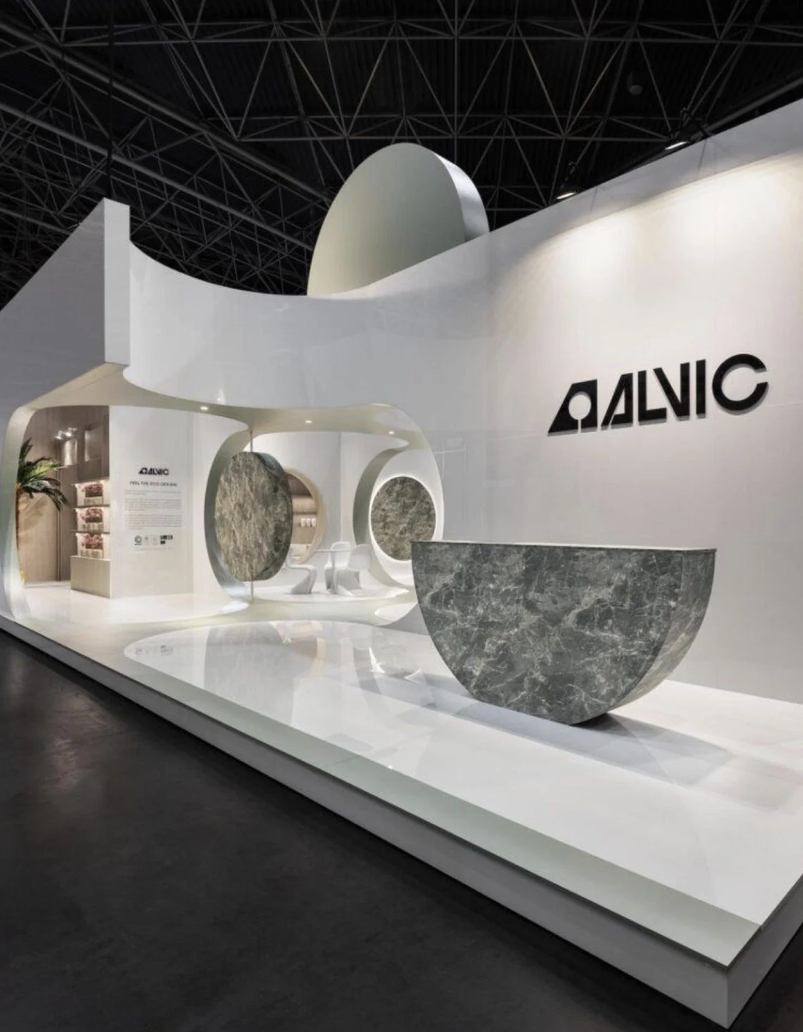 Trade show booth with white walls, abstract curved structures, and large stone-like bowls, displaying a sign that reads 'ALVIC' and additional information in the background.