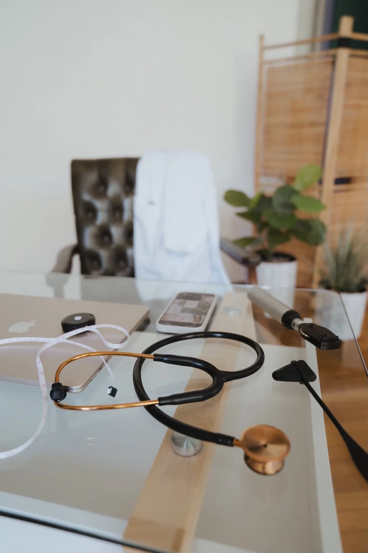 Medical office desk with stethoscope, smartphone, Apple MacBook, and medical tape measure, with a brown chair, green plants, and a wooden screen in the background.