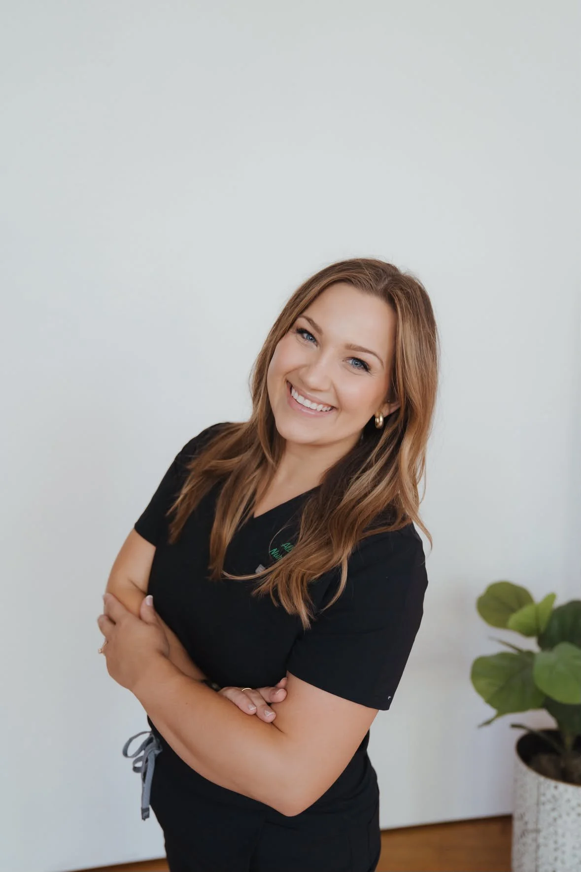 A woman with long, wavy, auburn hair smiling confidently with her arms crossed, wearing a black medical uniform, standing in a room with a plain white wall and green houseplant.