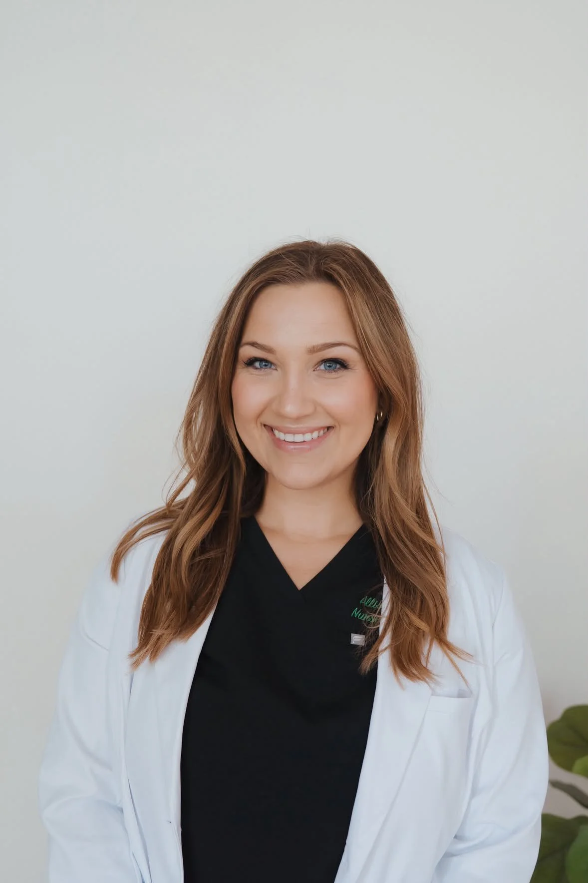 A young woman with long, wavy, auburn hair and blue eyes smiling, wearing a white coat over a black shirt, standing in front of a plain light-colored background with a small green plant on her right.
