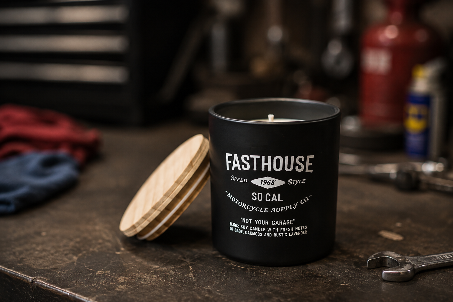 Black candle with white text and a wooden lid, placed on a workbench in a garage or workshop, with tools and supplies in the background.