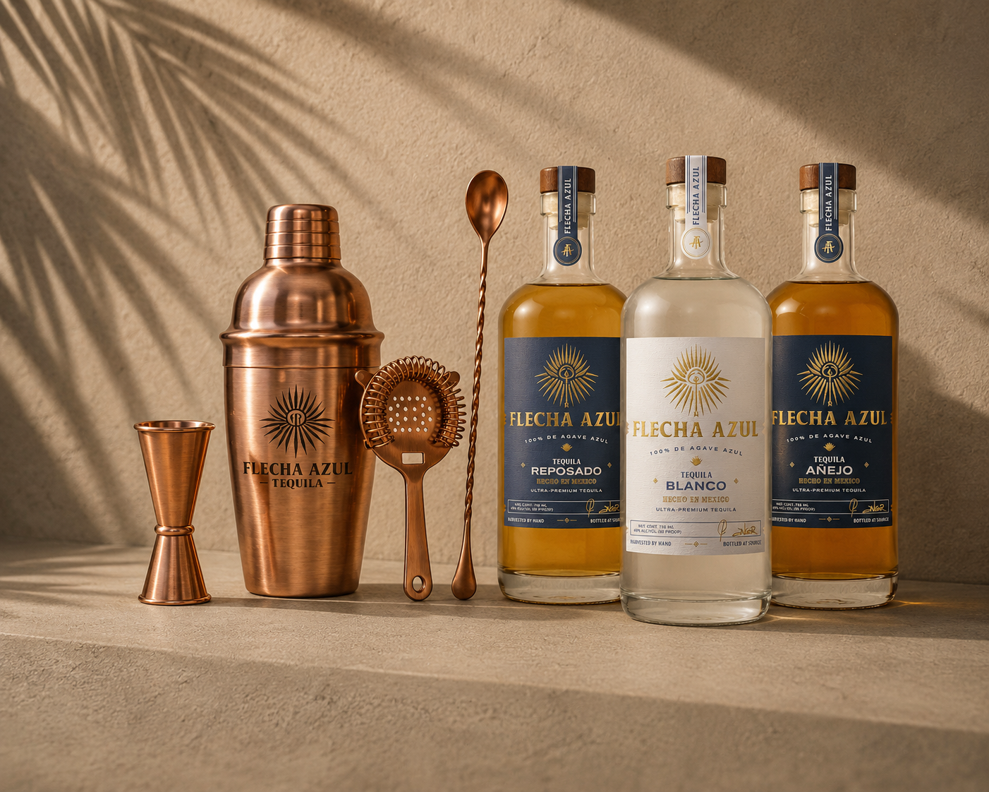 Set of three bottles of Flecha Azul tequila, a copper cocktail shaker, a copper jigger, a copper bar spoon, and a copper strainer on a beige surface with shadow and palm leaf shadow in the background.