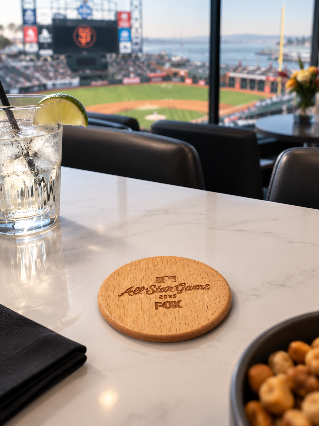 A glass of iced water with a lemon wedge on a white marble table, with a view of a baseball stadium through large windows in the background. A wooden coaster with 'All-Star Game 2025 FOX' engraved is on the table, along with a bowl of snacks and a bl