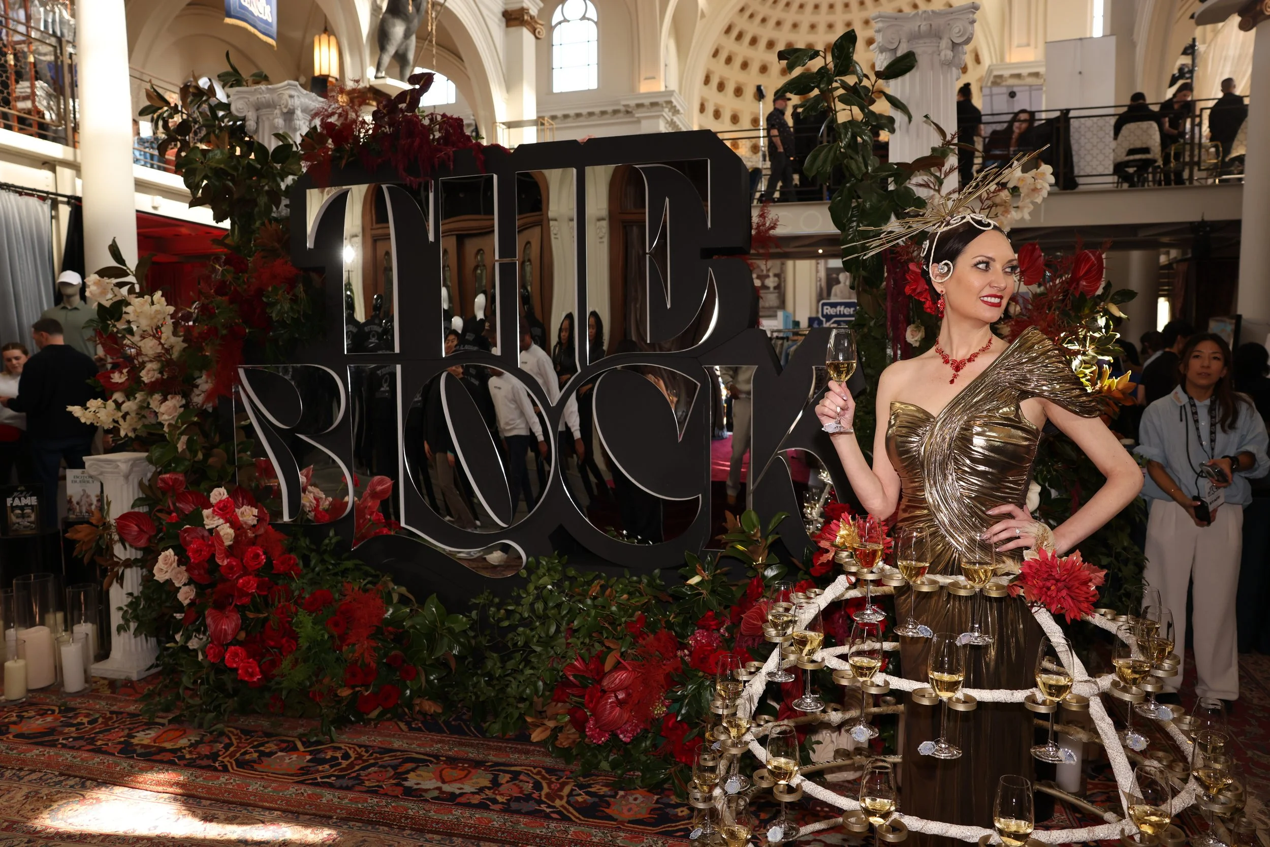 A woman in a metallic gold dress holding a glass of white wine, standing next to a floral display with red and white flowers, a large decorative sign that says 'LOVE', and a table filled with wine glasses in an indoor venue with a high, ornate ceilin