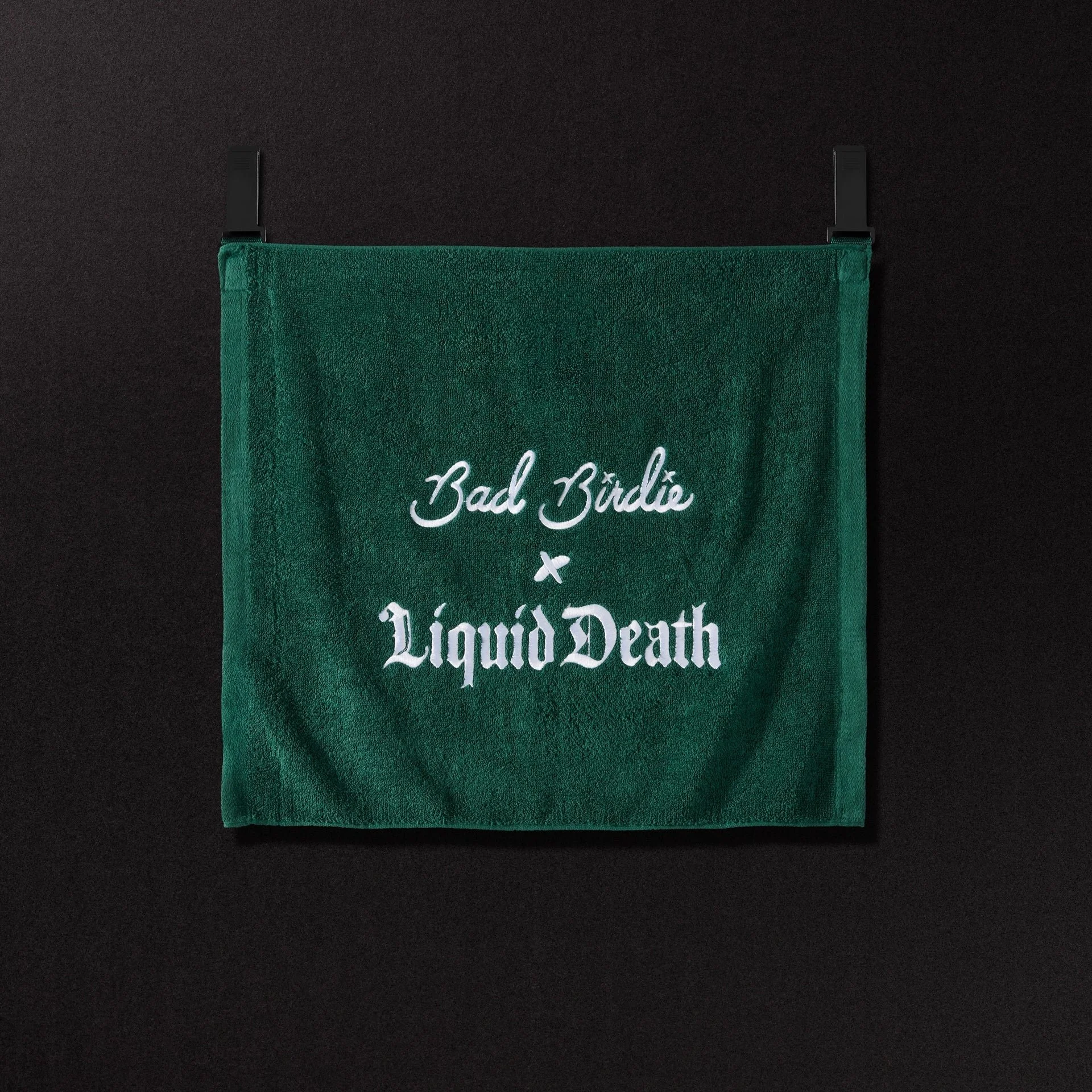 Close-up of a green cloth bag with white text reading 'Bad Biddie Liquid Death' against a black background.