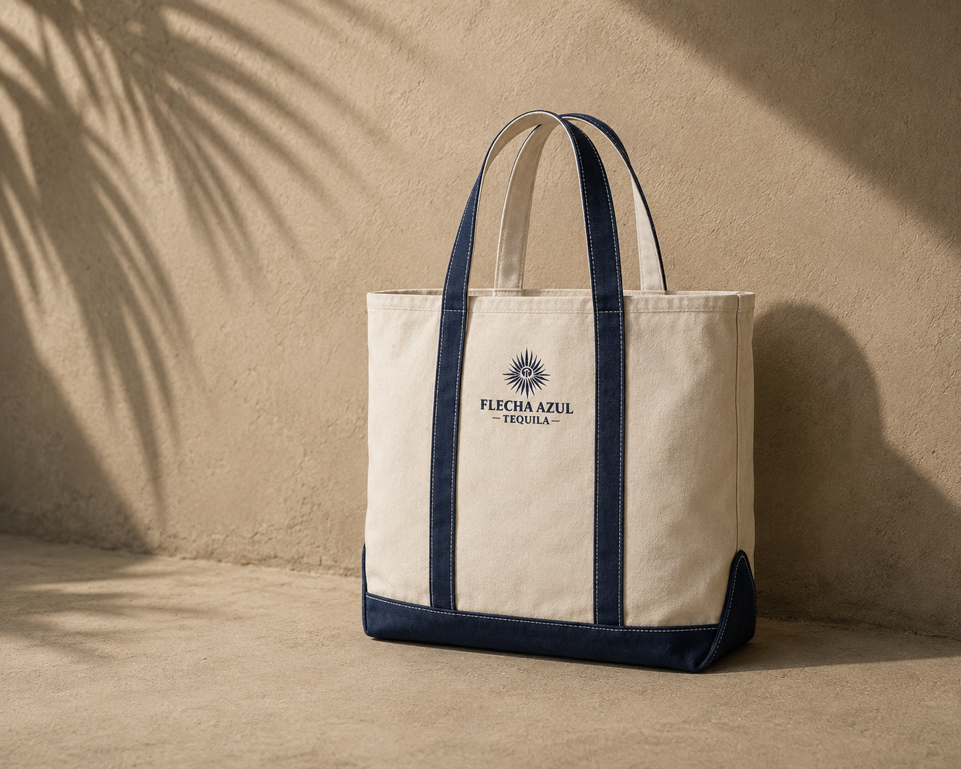 A beige tote bag with navy blue handles and trim, featuring a logo and the words 'FLECHA AZUL TEQUILA,' set against a sandy-colored background with shadows.