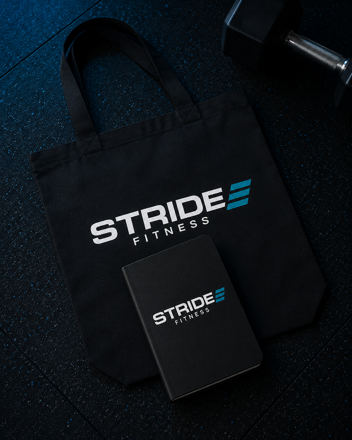A black gym tote bag, a notebook, and a water bottle are on a dark speckled gym floor. The tote bag and notebook display the 'Stride Fitness' logo.