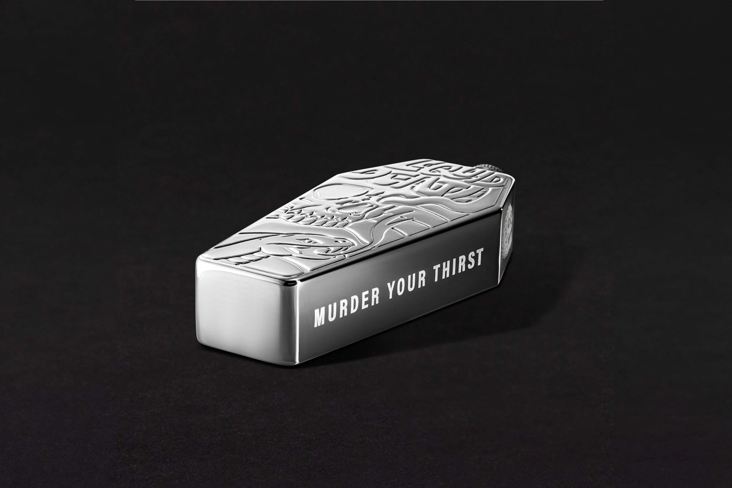 A silver box with intricate engraved designs on the top, featuring the phrase "Murder Your Thirst" in black letters on the side, set against a black background.
