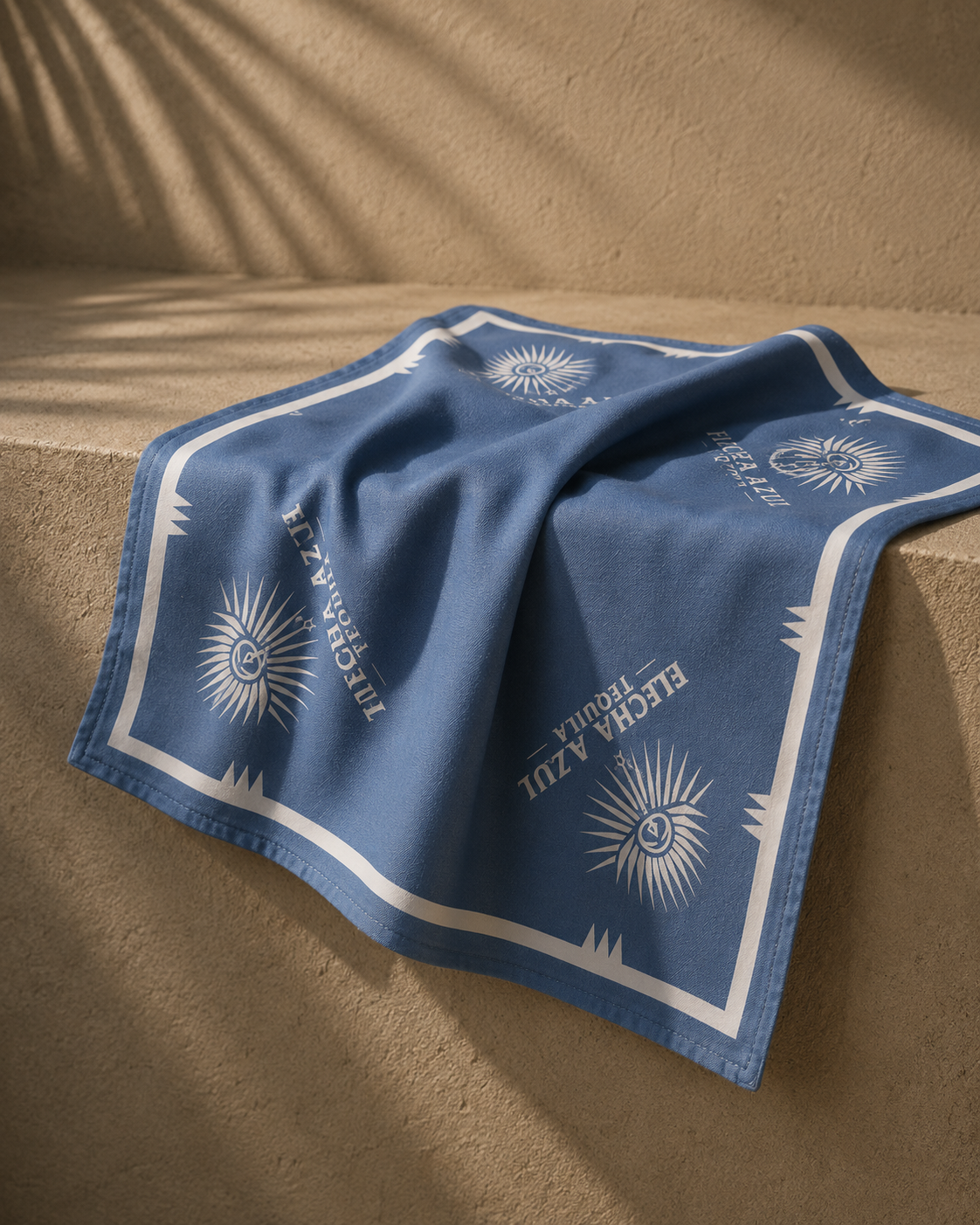 A blue handkerchief with white logos and text, draped over a textured beige surface, with shadows cast by nearby palm leaves.