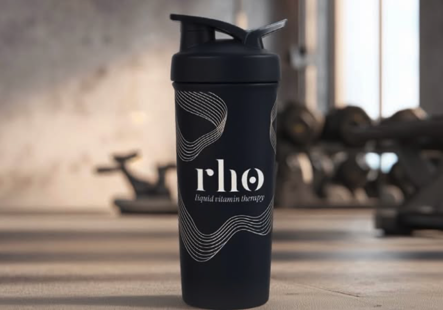 A black water bottle with the logo 'flo' and the text 'liquid vitamin therapy' on it, placed on a wooden gym bench with dumbbells in the background.